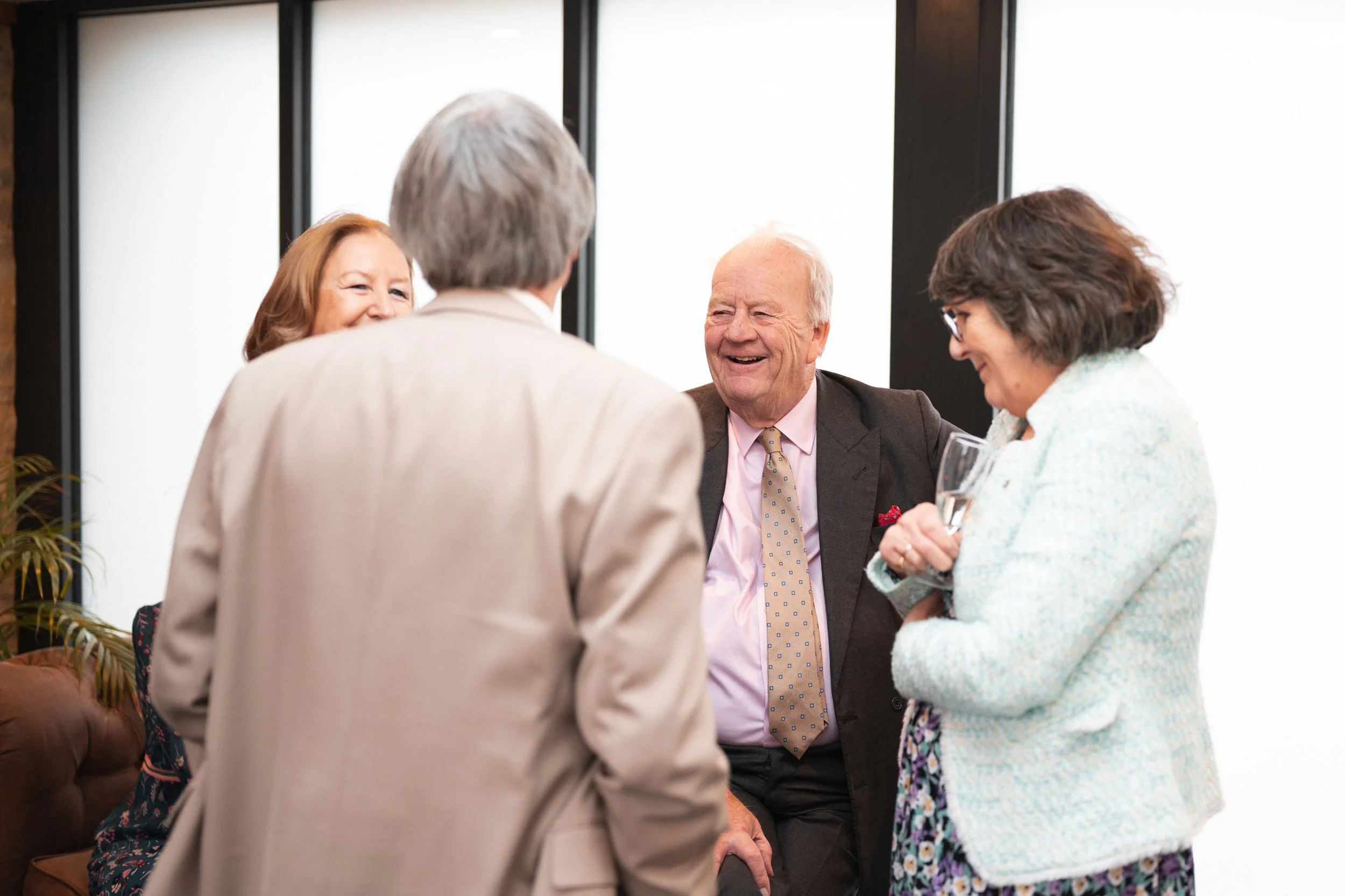Four elderly people, two men and two women, are gathered in a semi-circle indoors, engaged in conversation and smiling. One woman is holding a glass of champagne, while the others are listening and talking.