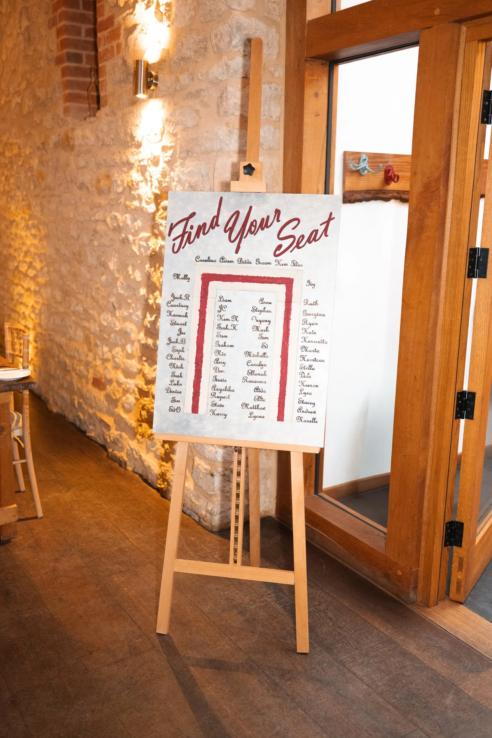 Event seating chart titled 'Find Your Seat' with a list of names and a red outline of a doorway, displayed on an easel inside a rustic venue.