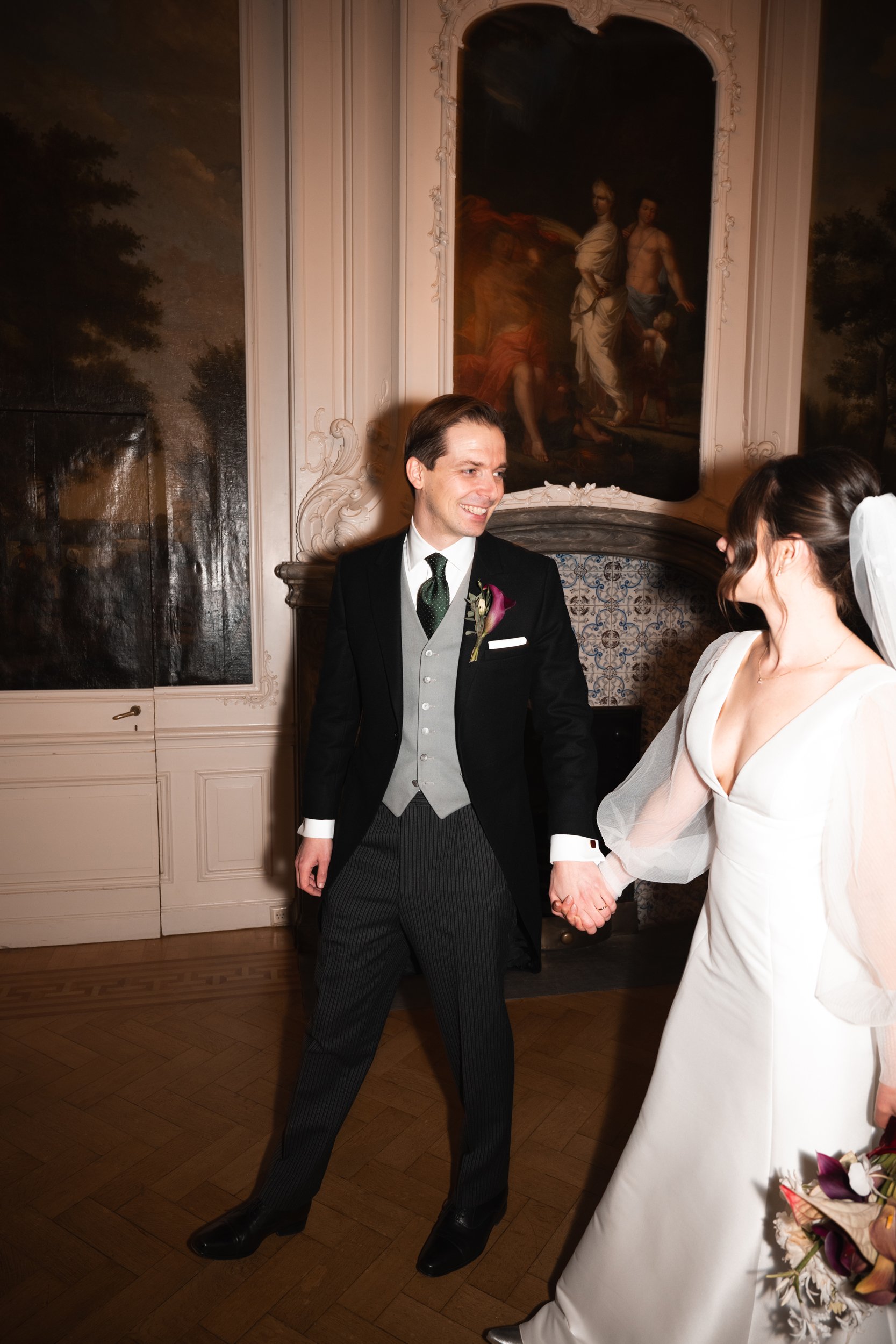 A groom and bride holding hands in a room with ornate walls and classical paintings, sharing a joyful moment at their wedding.