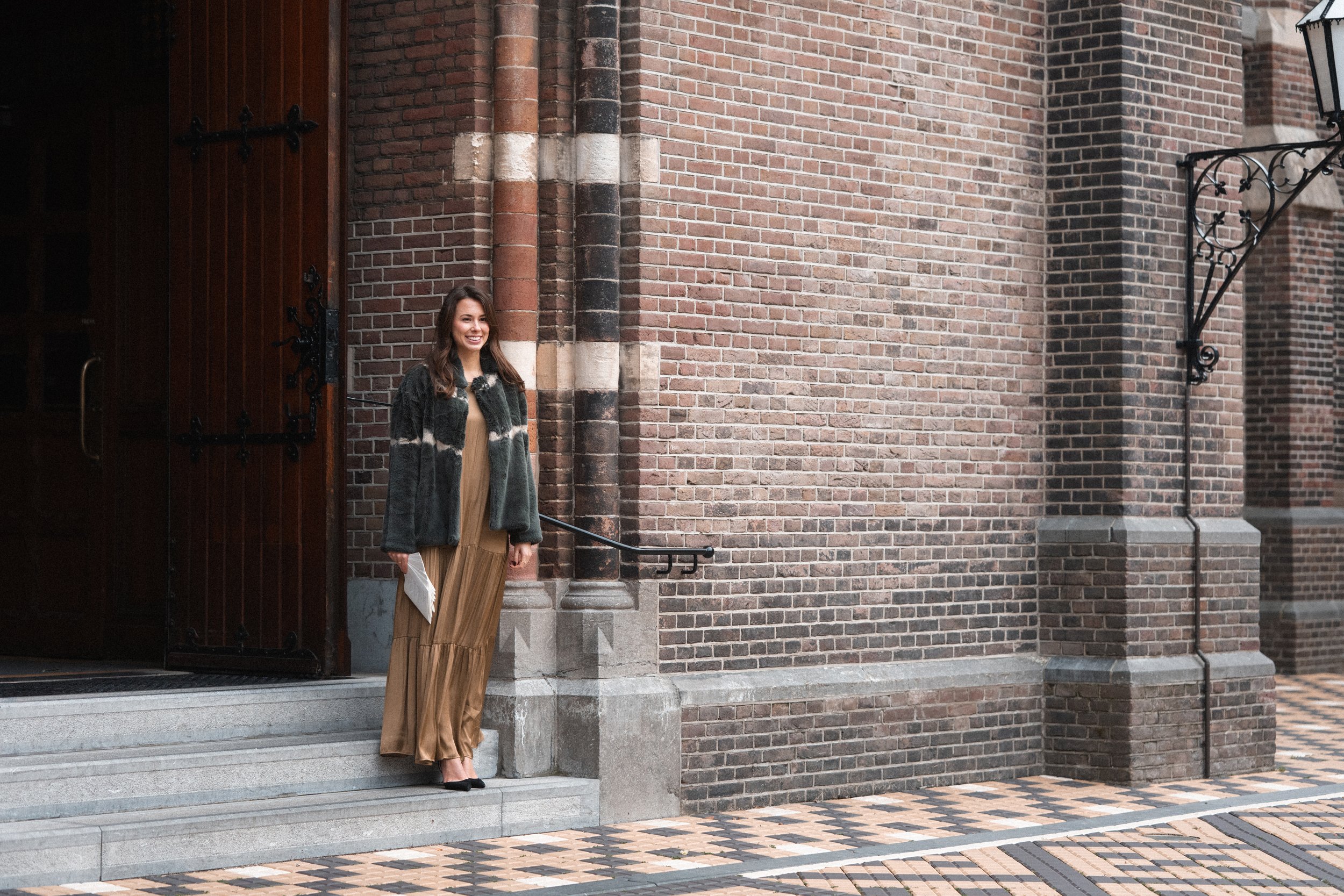 A woman standing on grey stone stairs outside a brick building, holding a white item, smiling, wearing a beige dress, black shoes, and a grey jacket.