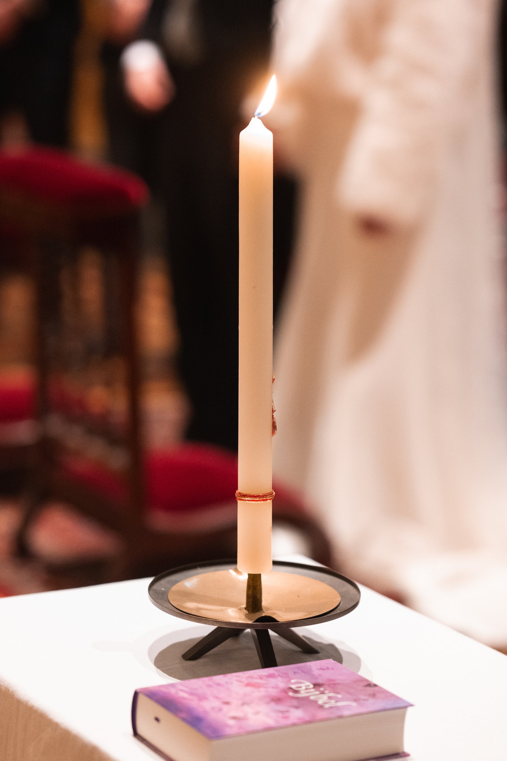 A white lit candle on a candle holder with book nearby, on a table during a religious ceremony.