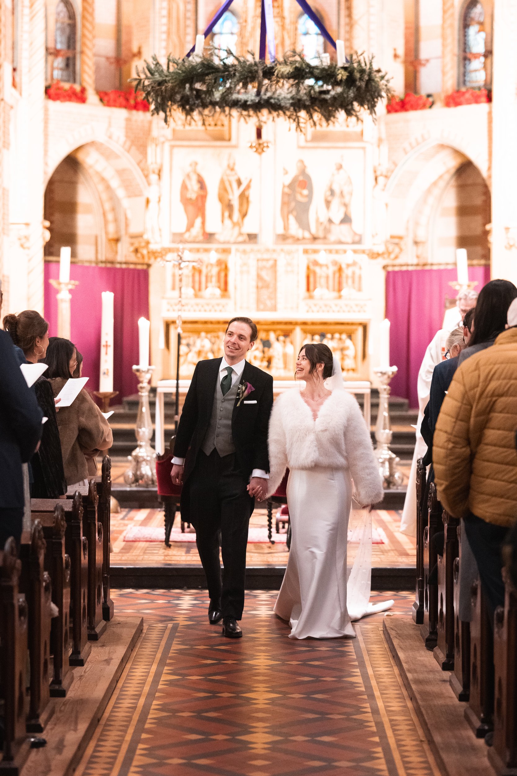 A newlywed couple walking hand in hand down the aisle of a church, surrounded by guests, with an ornate altar and religious paintings in the background.