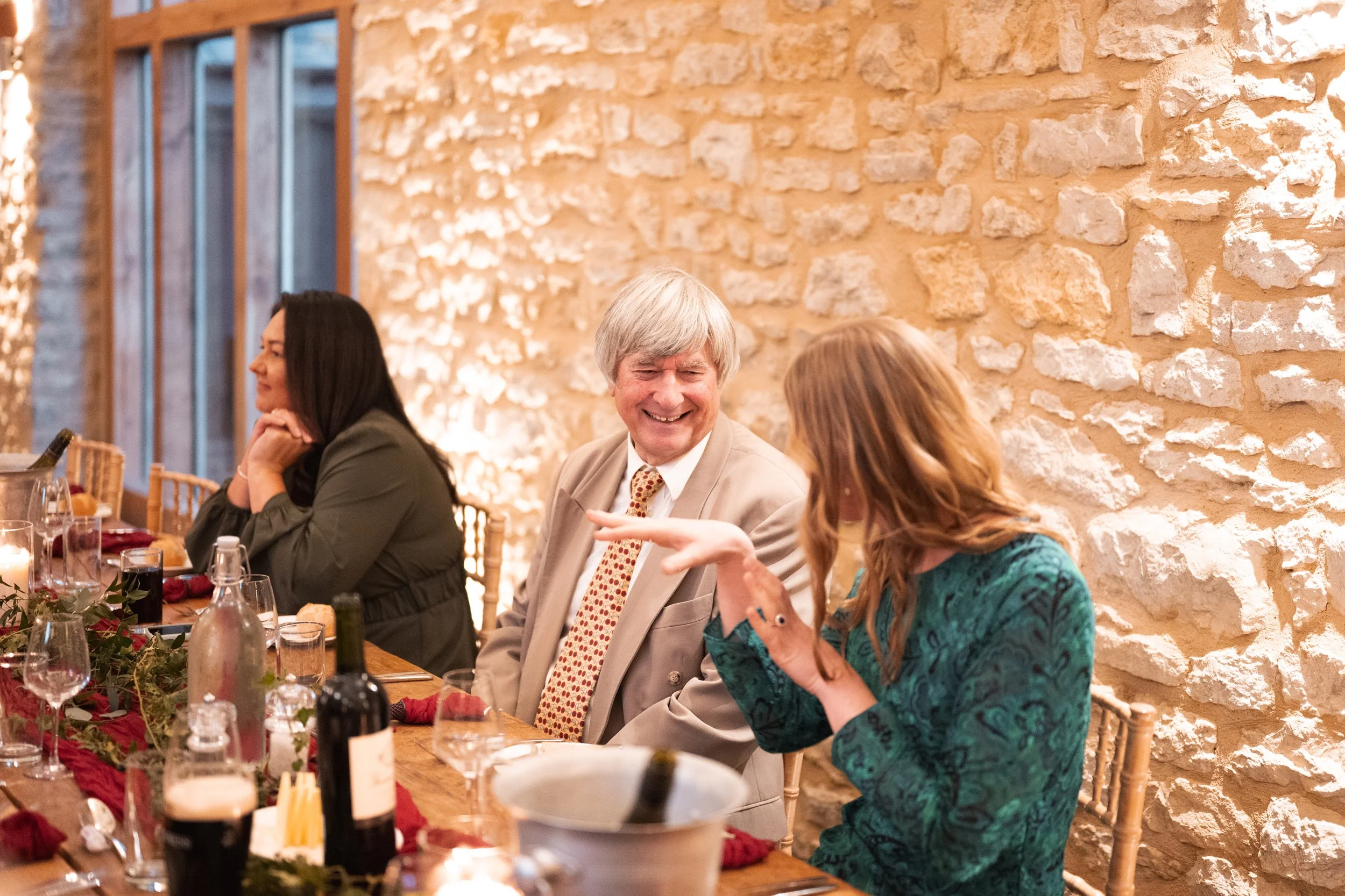 People sitting at a long dining table, engaging in conversation, in a well-lit room with a brick wall background.