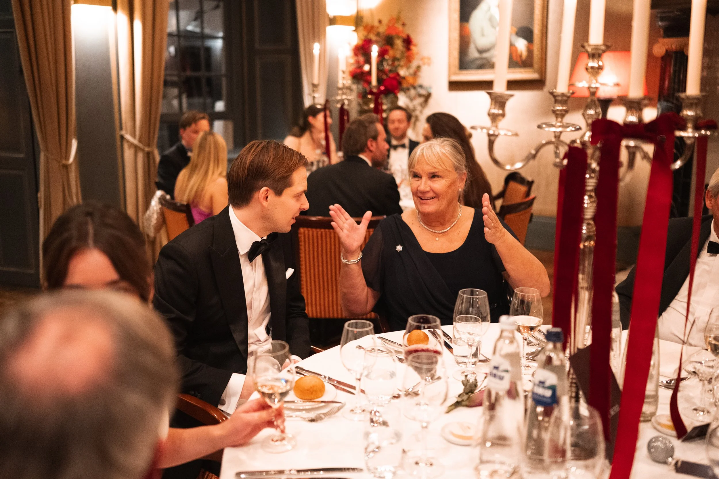 People sitting at a dinner table, engaging in conversation, in an elegant banquet setting with chandeliers, candles, and floral centerpieces.