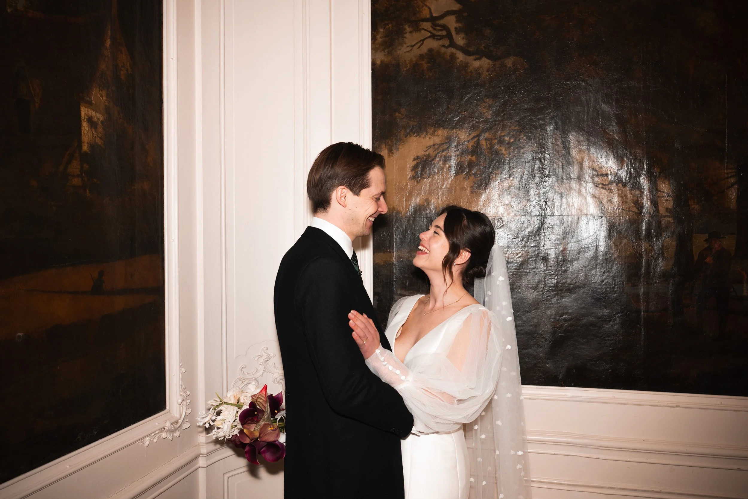A bride and groom looking at each other and smiling indoors, with dark wall art in the background. The bride is wearing a white wedding dress and veil, and the groom is in a black suit.