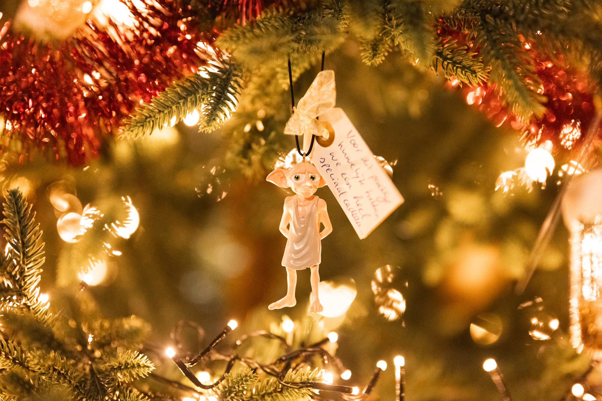 A Christmas tree ornament of a small creature with large eyes and large ears, wearing a simple dress, hanging from a decorated Christmas tree with warm lights and greenery, along with a handwritten note.