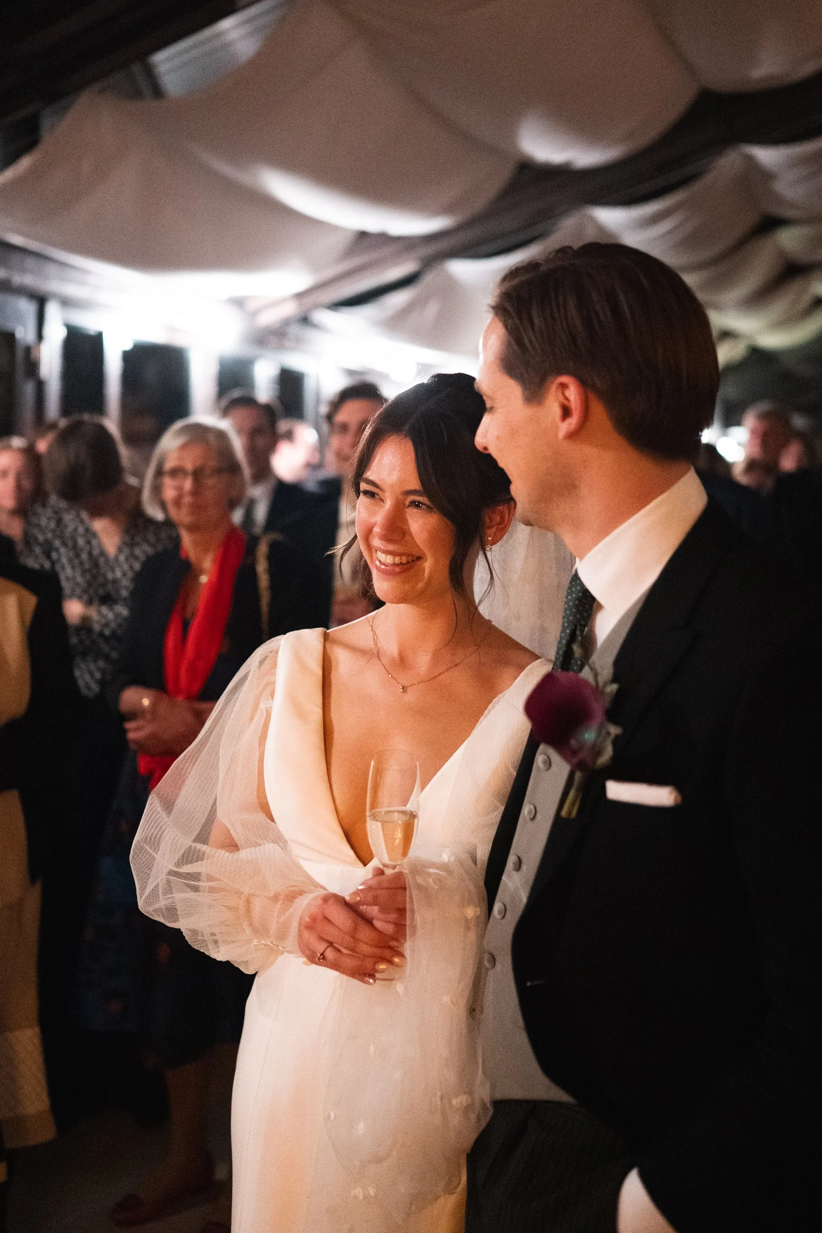 A bride and groom smiling at each other at their wedding reception, with guests in the background.