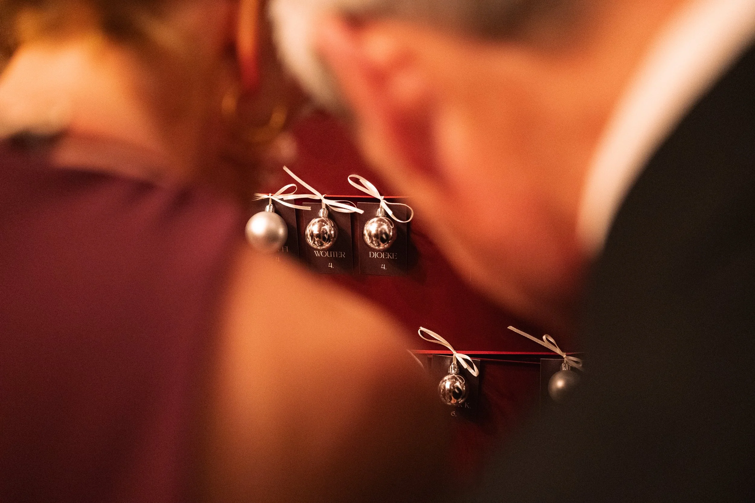 Close-up of wedding ring charms hanging on a red background, with the names 'Wouter' and 'Djoeke' visible on some tags, surrounded by blurred figures of two women leaning in close to each other.