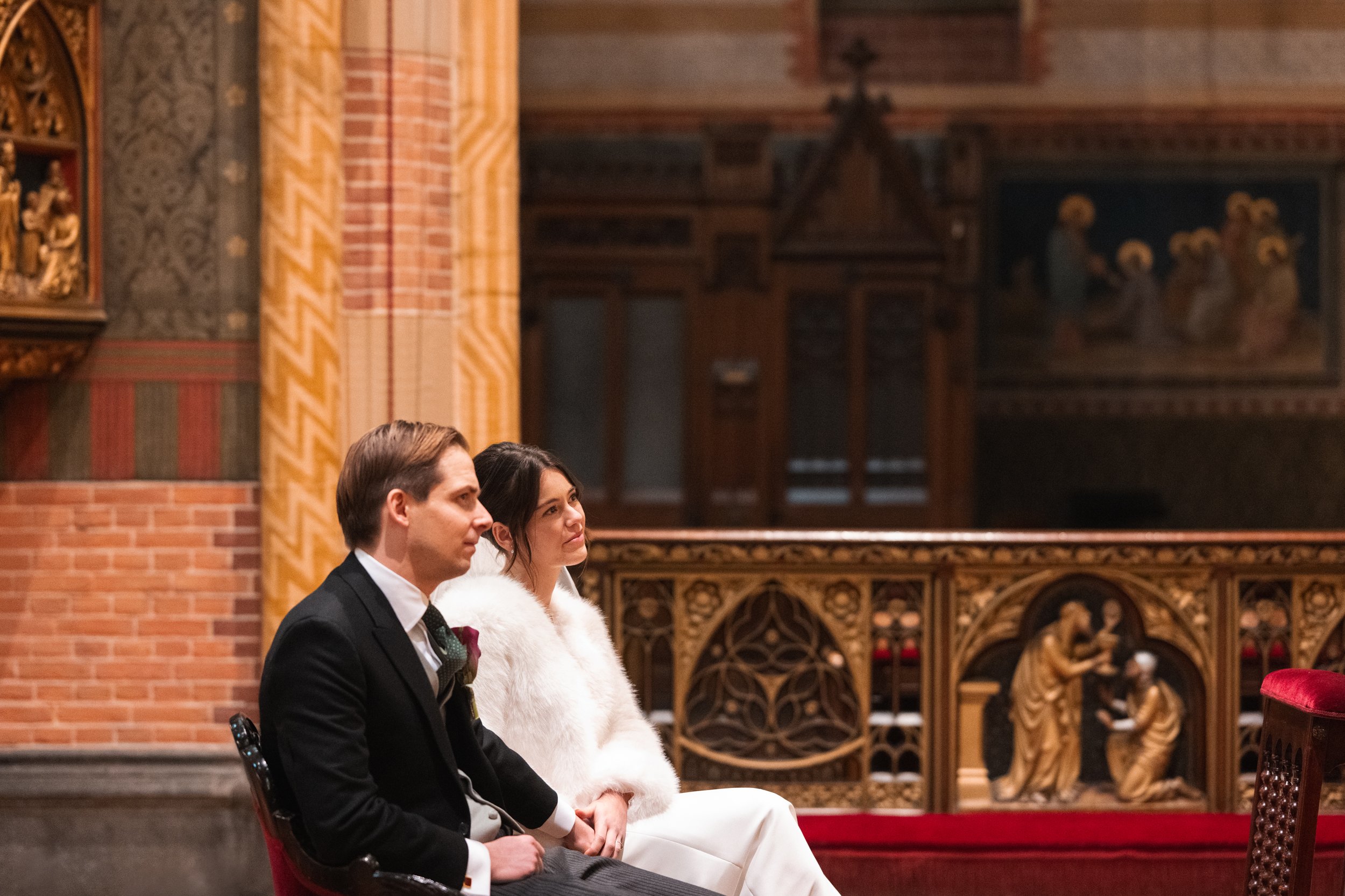 A man and woman sitting inside a church or cathedral, dressed formally. The man is in a black suit, and the woman is in a white dress with a white fur coat. They are seated on dark wooden chairs with red upholstery, with an ornate wooden railing and 