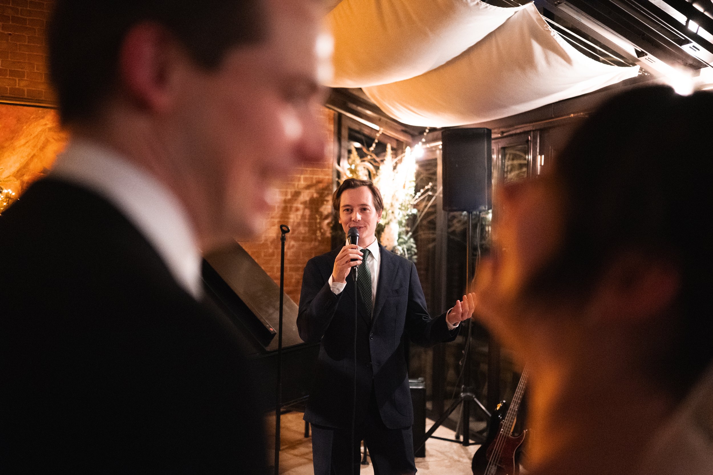 Person in a suit speaking into a microphone at an event with blurred foreground people, indoor setting with warm lighting, brick wall, speaker, microphone stand, guitar, and floral or decorative arrangements in the background.