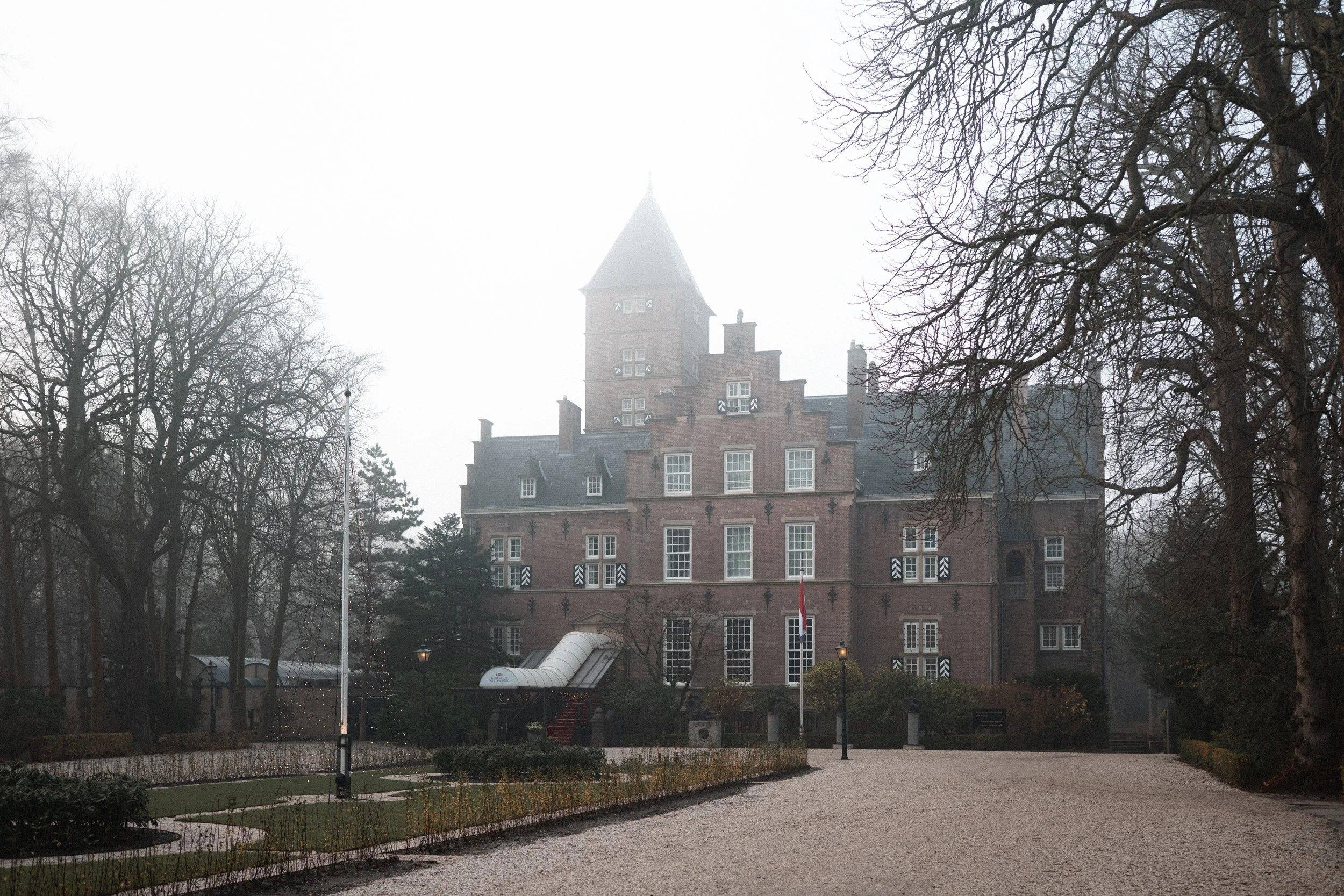 An old brick castle with multiple stories and a tall tower, surrounded by leafless trees on a foggy day.