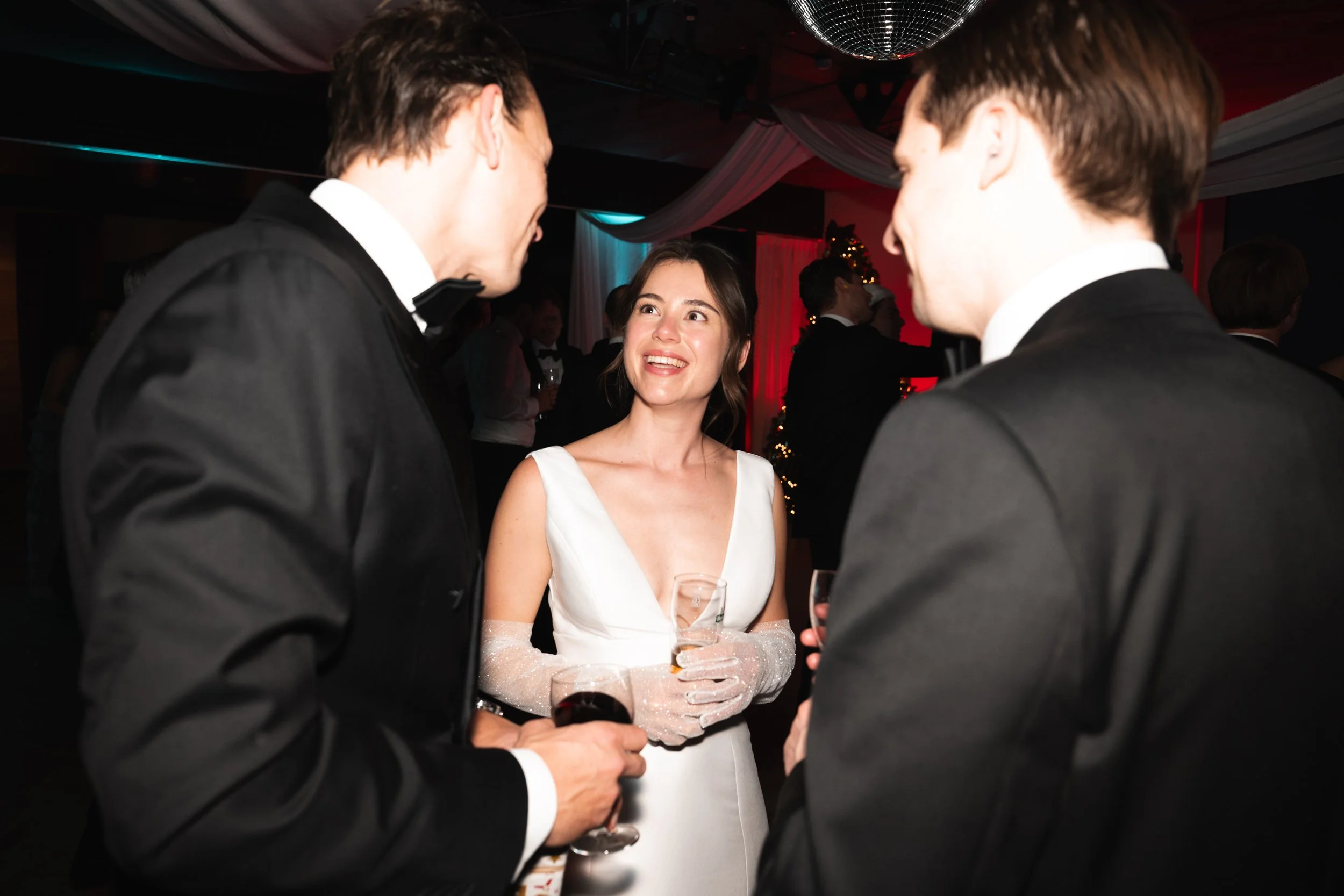 A woman in a white dress smiling and talking with two men in tuxedos at a formal event.