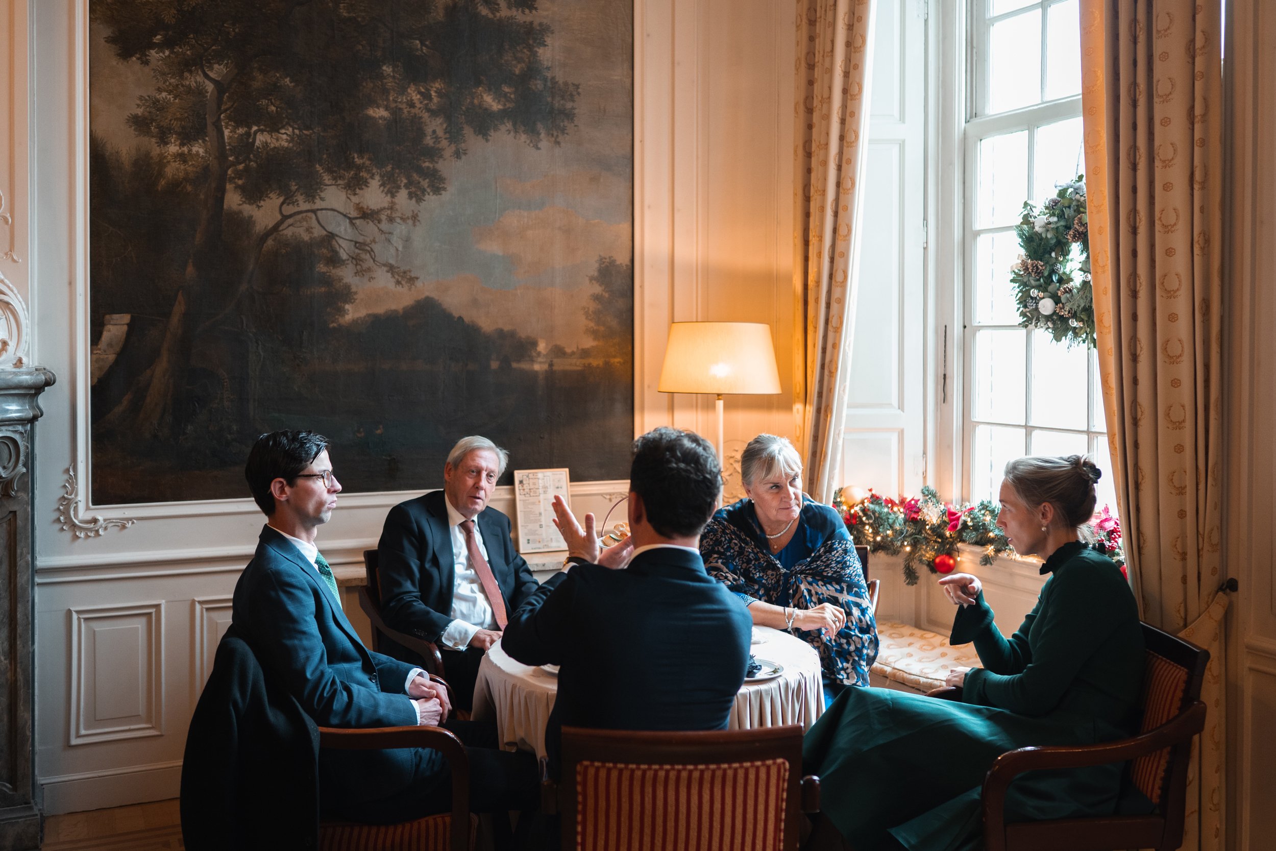 Five adults seated around a round table having a conversation in a festively decorated room with a Christmas wreath on the window and holiday decor.