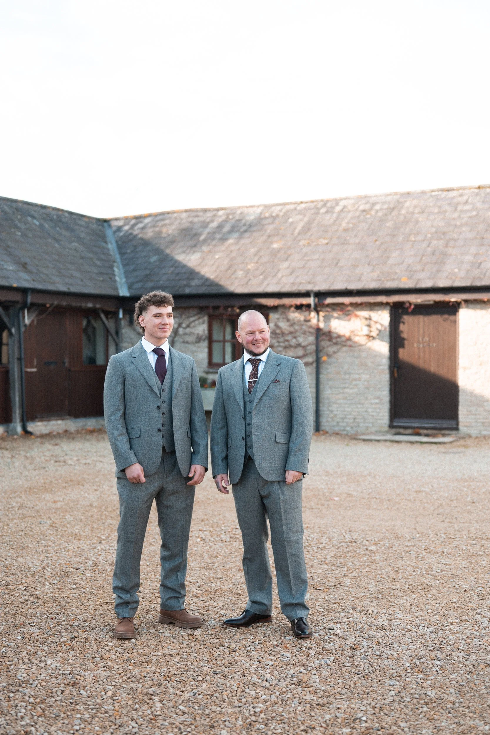 Two men in grey suits standing outdoors, smiling in front of a barn or rustic building.