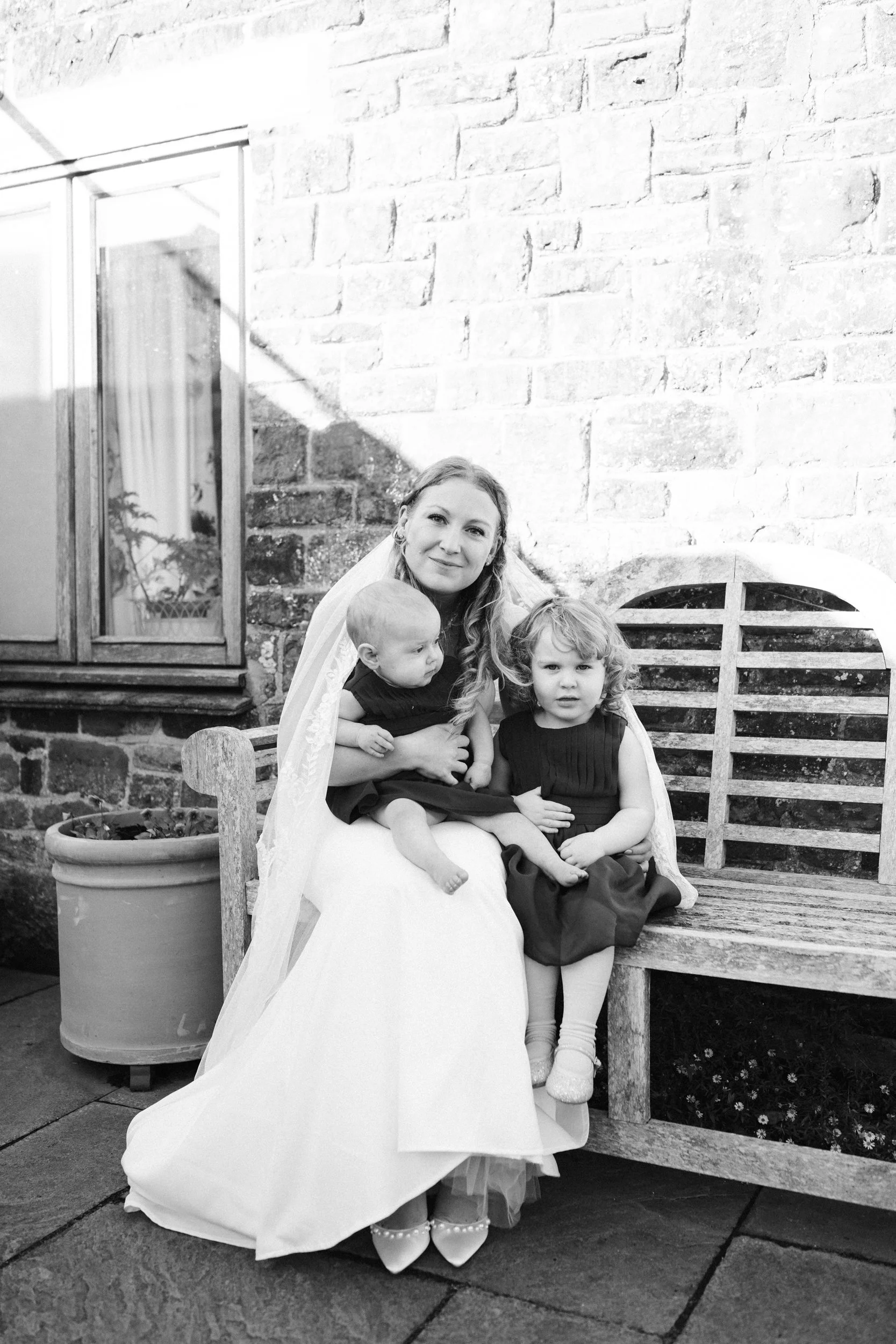 A woman in a wedding dress sitting on a wooden bench outdoors, holding two young girls in dark dresses on her lap, in front of a brick wall with a window and potted plant nearby.