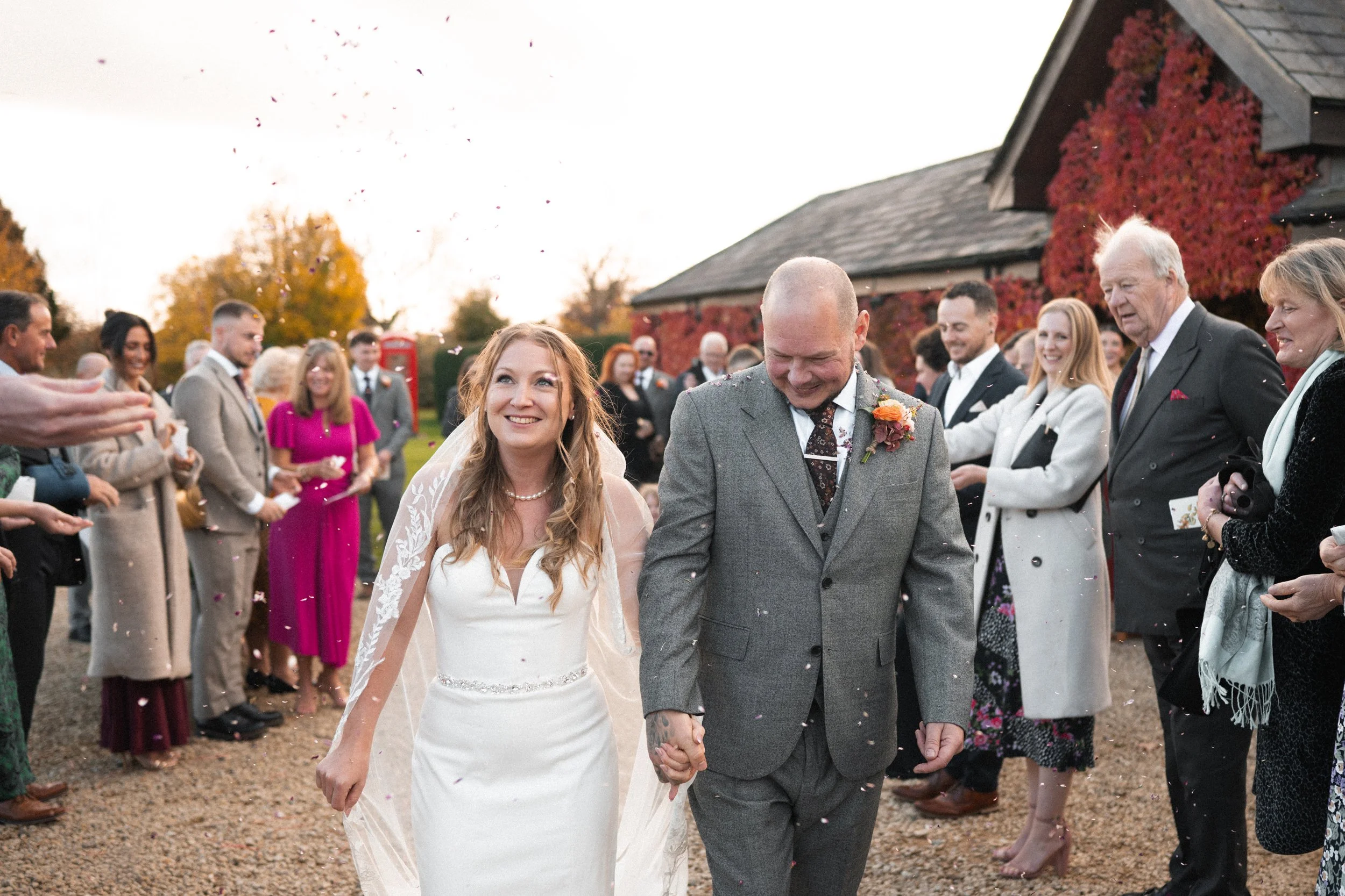 A bride and groom walking down a gravel path holding hands, smiling, surrounded by guests celebrating their wedding outdoors, autumn trees in background.