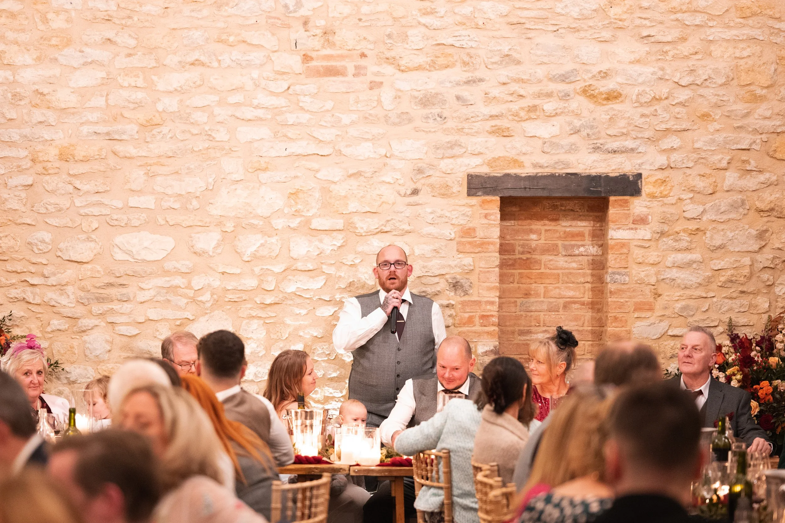 A man in a gray vest and tie giving a speech at a wedding reception, surrounded by seated guests at decorated tables, inside a rustic stone-walled venue.