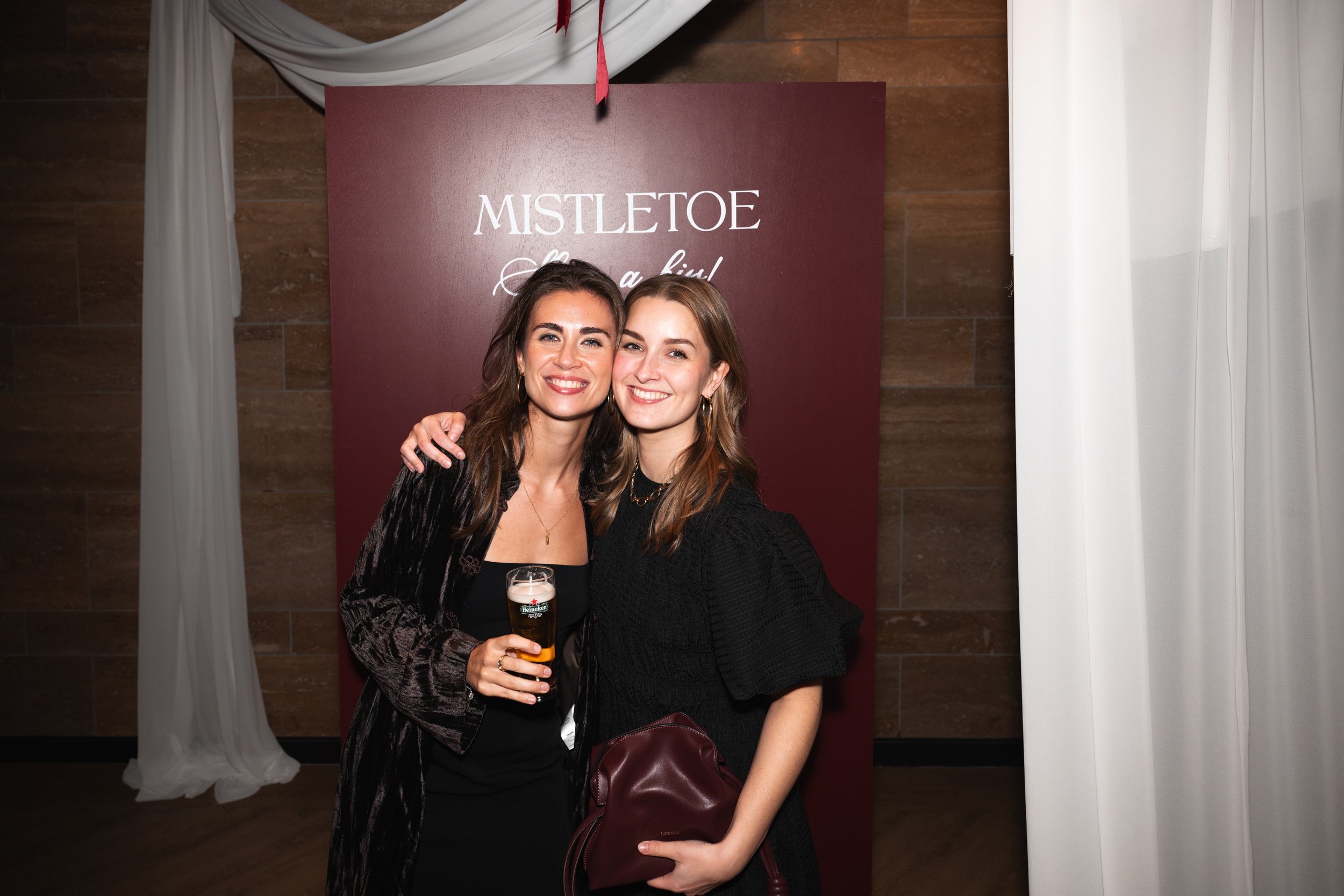 Two women smiling and hugging at a party, one holding a glass of beer, standing in front of a maroon backdrop with white text that reads 'MISTLETOE' and some white curtains on the sides.