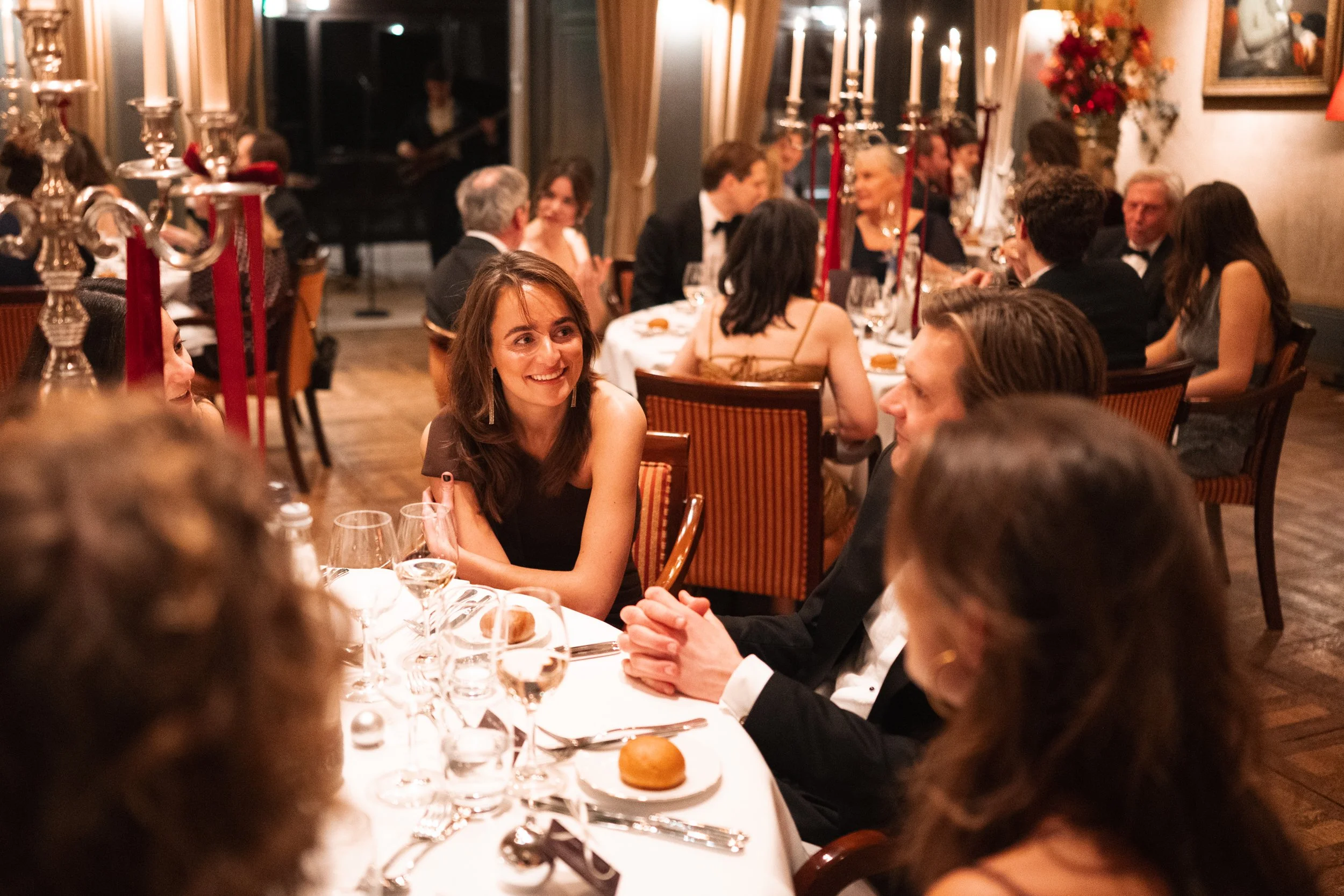 People attending a formal dinner party in an elegant dining room with candles and floral decorations, engaging in conversation and enjoying the event.