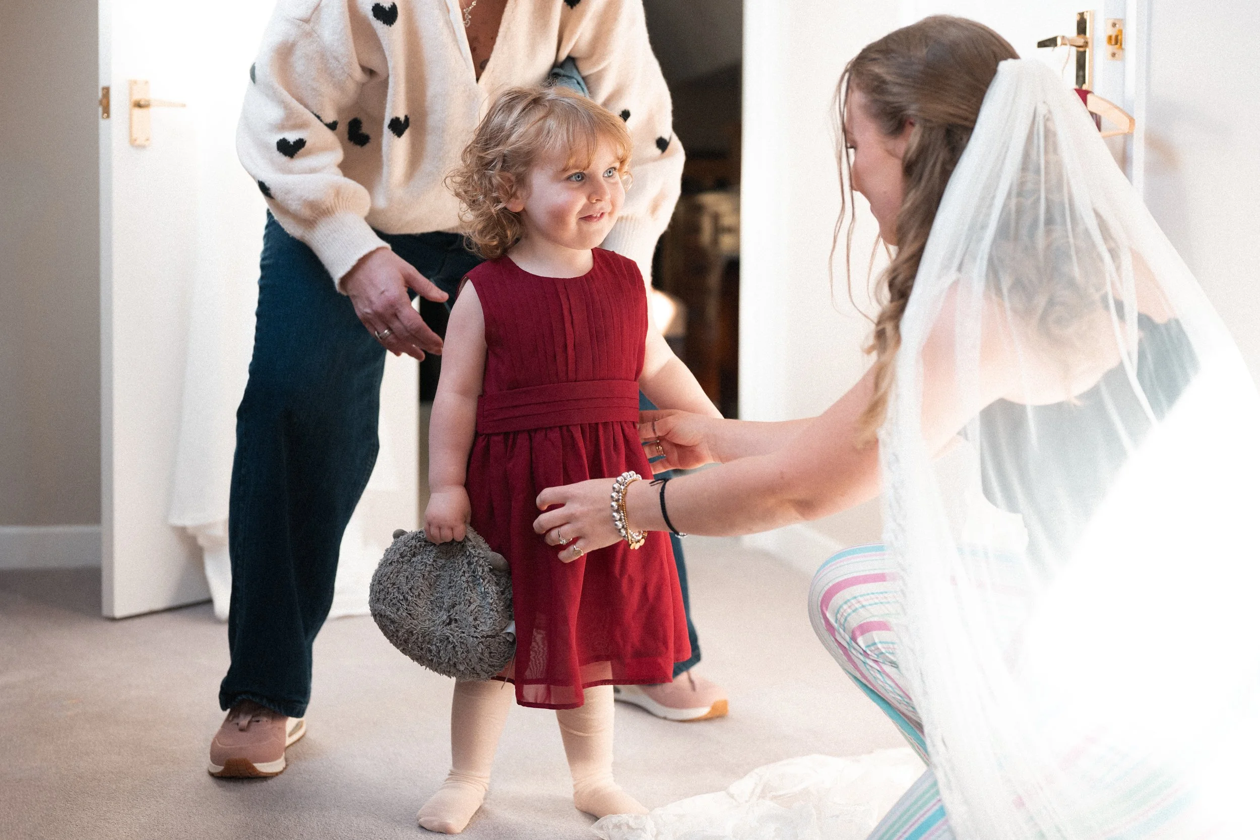 A young girl in a red dress looks at a woman dressed as a bride with a veil, who is kneeling and adjusting the girl's dress. An older woman stands behind them, watching.