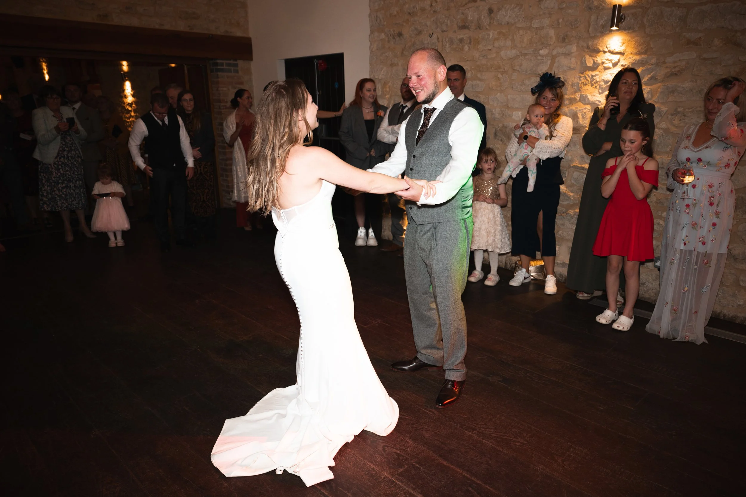 A bride and groom are dancing at their wedding reception, surrounded by guests watching and smiling.