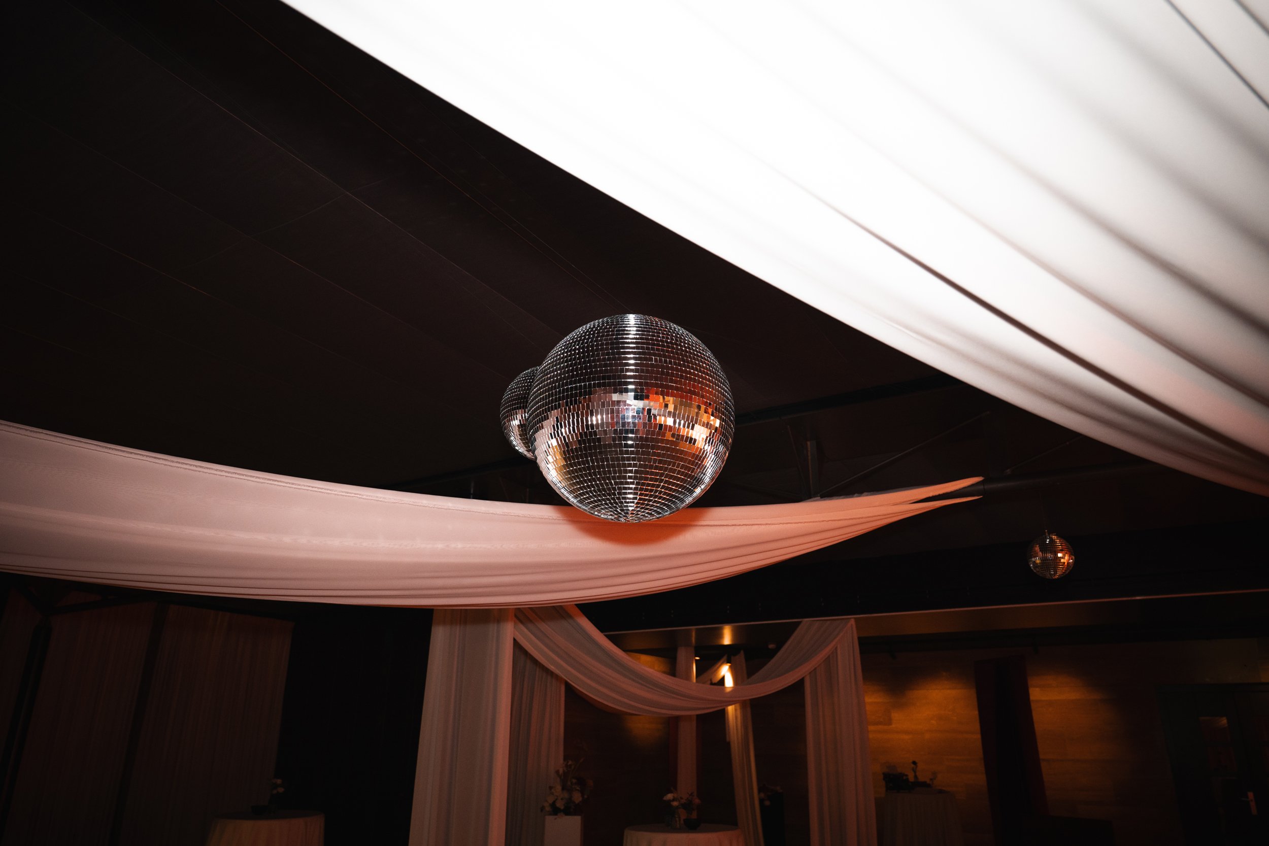 Mirror ball disco balls hanging from the ceiling with draped white fabric in the background of a decorated event space.
