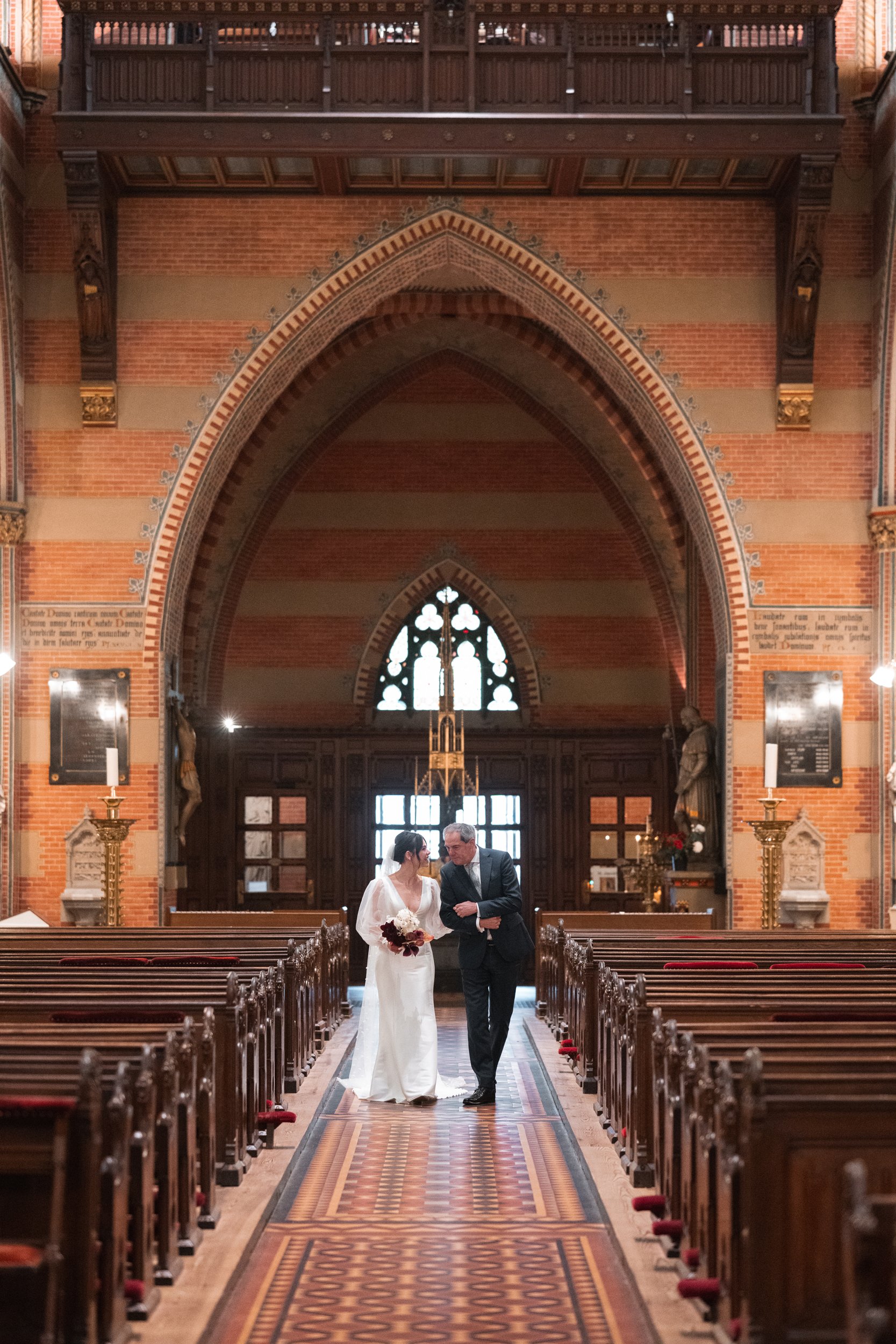 Bride and groom walk down the aisle inside a historic church with ornate architecture, stained glass windows, and wooden pews.