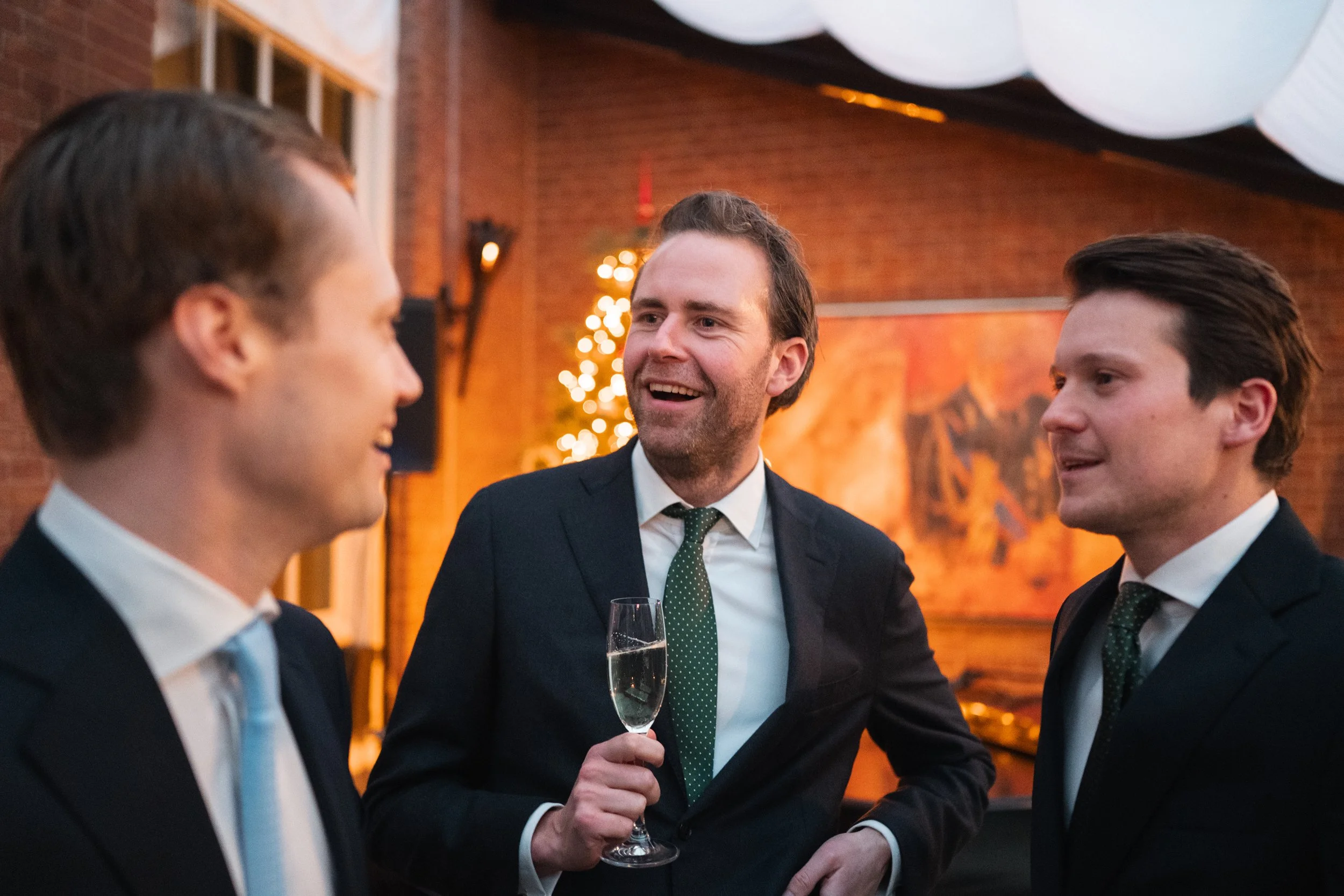 Three men in suits having a conversation at a social gathering, one holding a glass of champagne, in a warmly lit indoor venue decorated with a Christmas tree and artwork on brick walls.