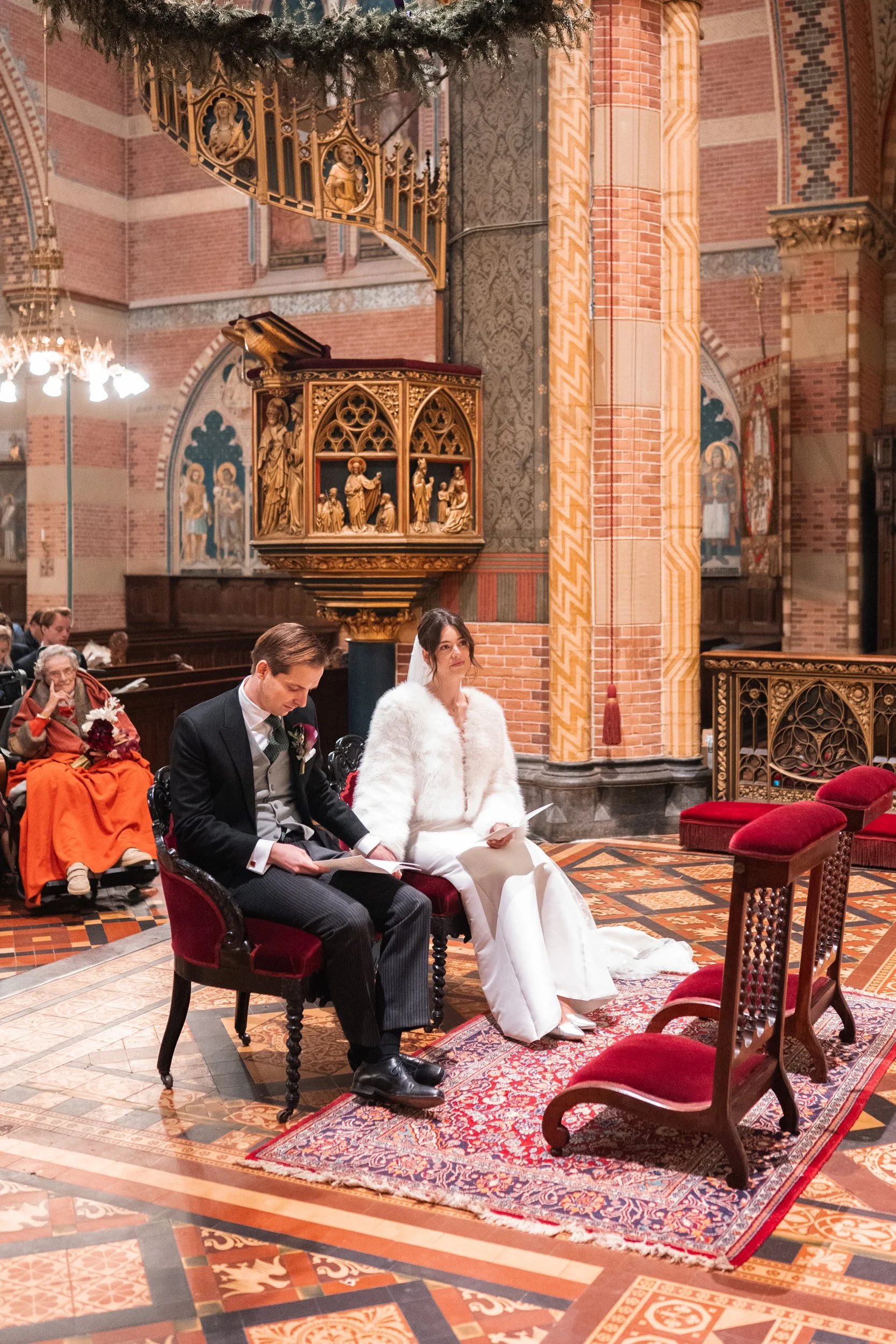 A bride and groom sitting on red velvet chairs during a wedding ceremony inside a church. The bride is wearing a white gown and a white fur shawl, while the groom is dressed in a dark suit with a vest and tie. An older woman dressed in orange is seat