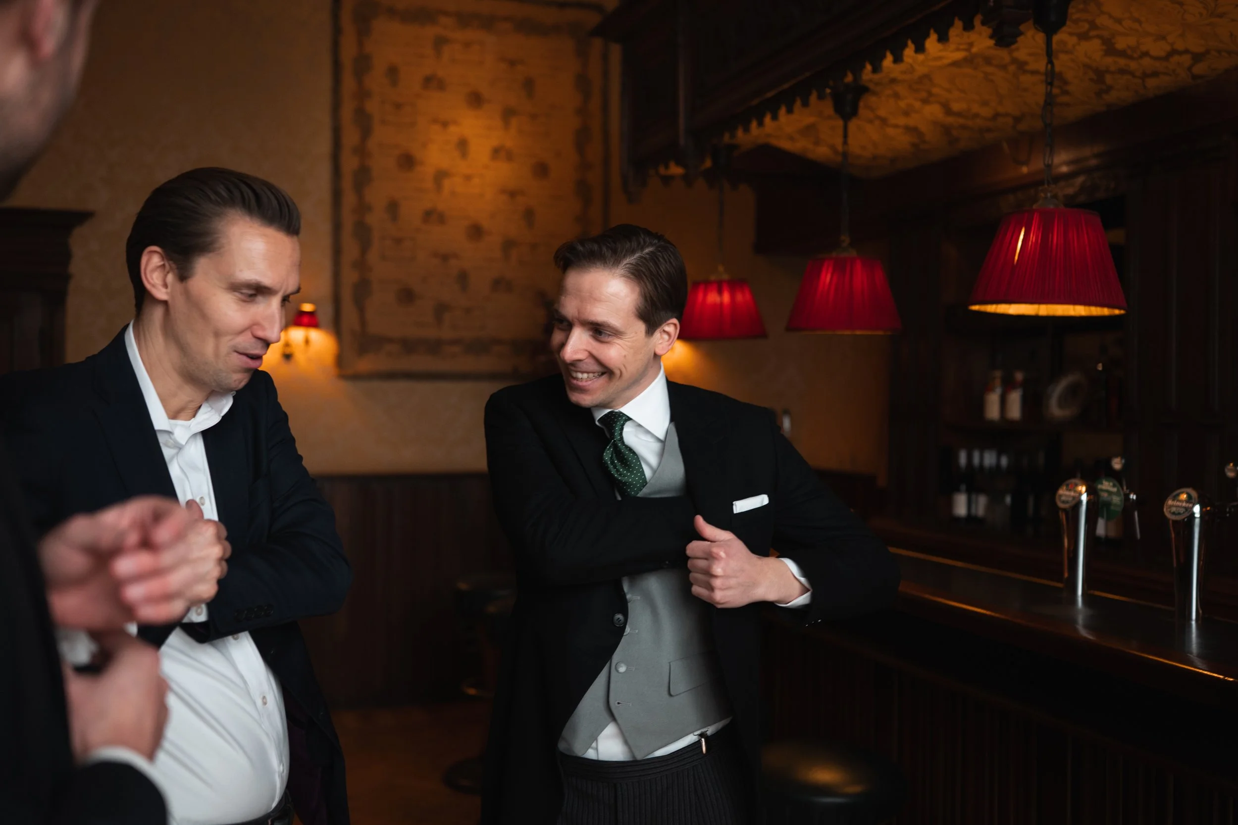 Two men in formal attire smiling and conversing inside a dimly-lit bar or restaurant with red lampshades hanging from the ceiling.