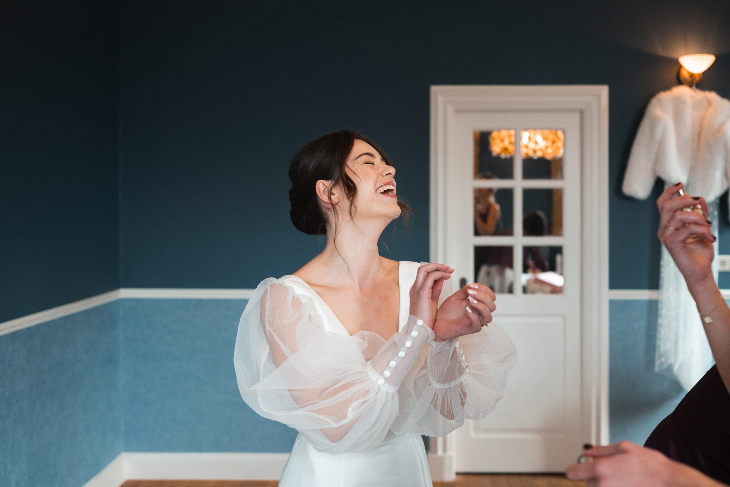 A woman in a white dress with sheer, puffy sleeves laughs joyfully in a room with dark blue walls and a mirror reflecting her and another person.