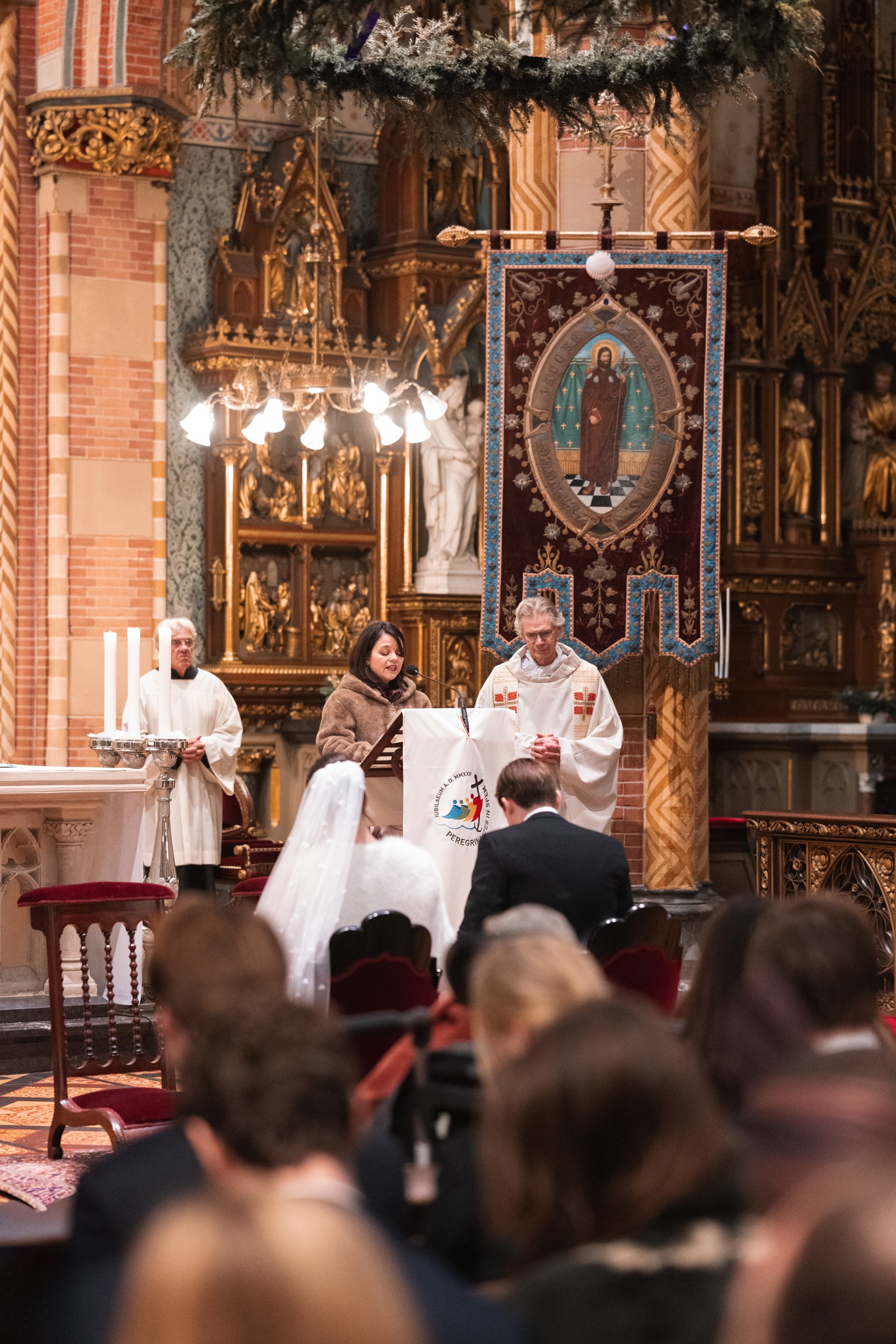 A church service with a priest, a woman, and a man at the altar, surrounded by church members, inside a decorated Catholic church with gold accents and religious icons.