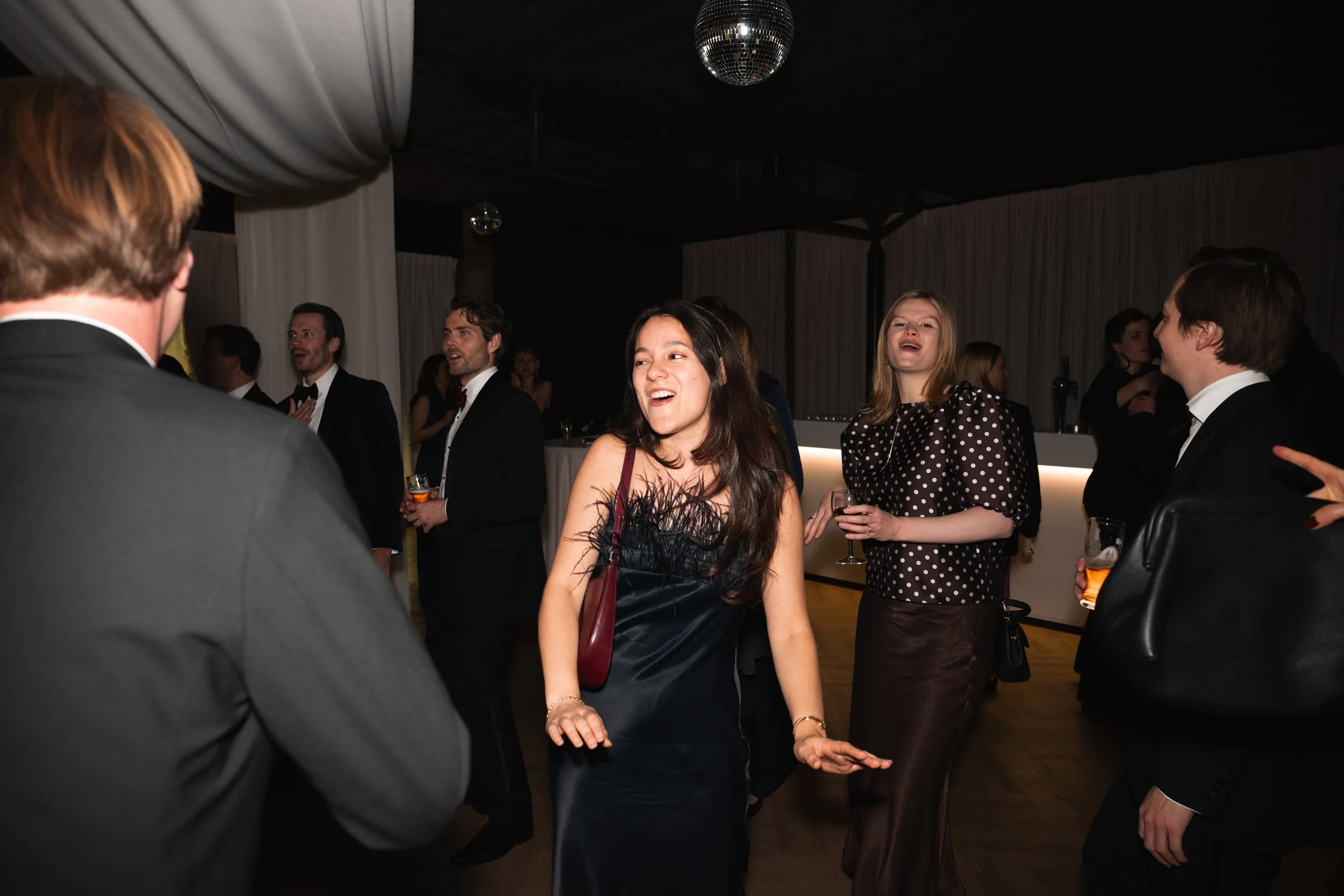 People dancing and socializing at a formal event or party in a dimly lit venue with white drapery and a disco ball.