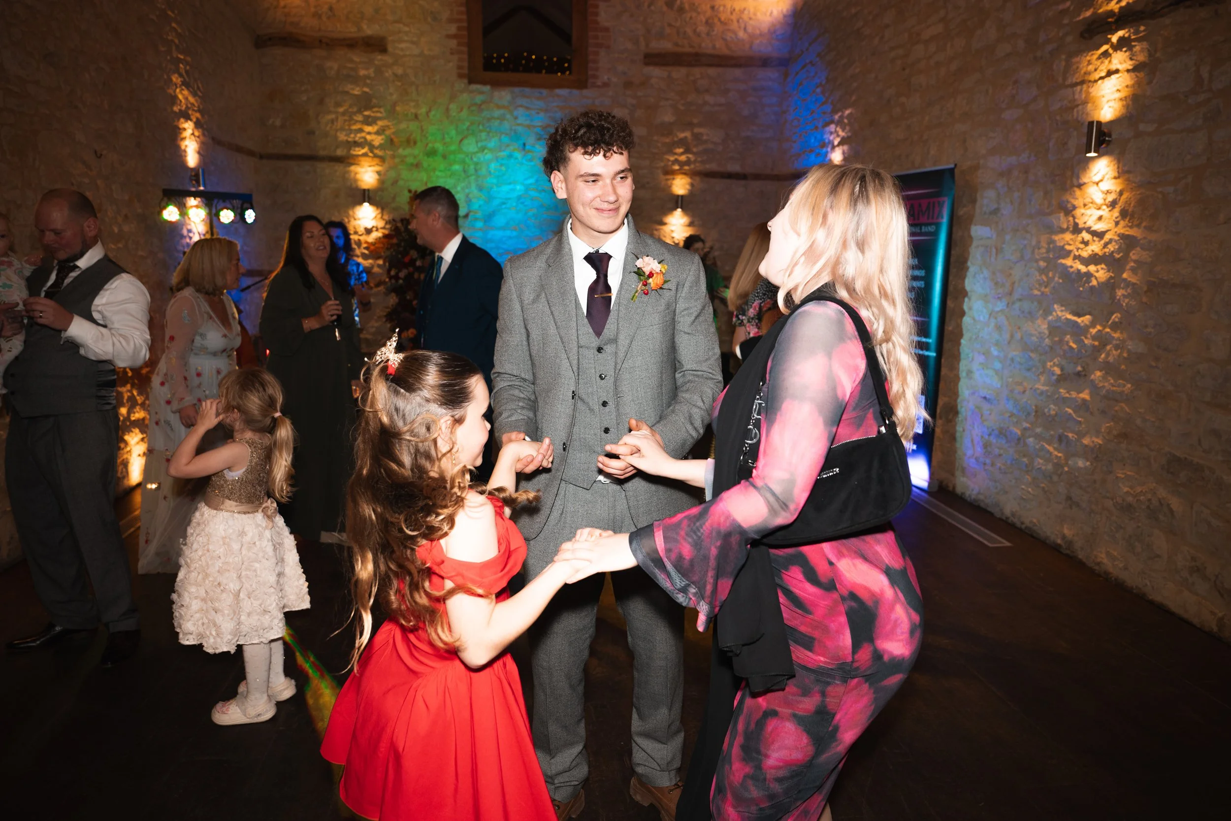 A group of people dancing and socializing at a party or wedding reception in a rustic stone-walled venue with colorful lighting. Two children and three adults are prominent, with a young man in a gray suit and a woman in a pink and black dress holdin