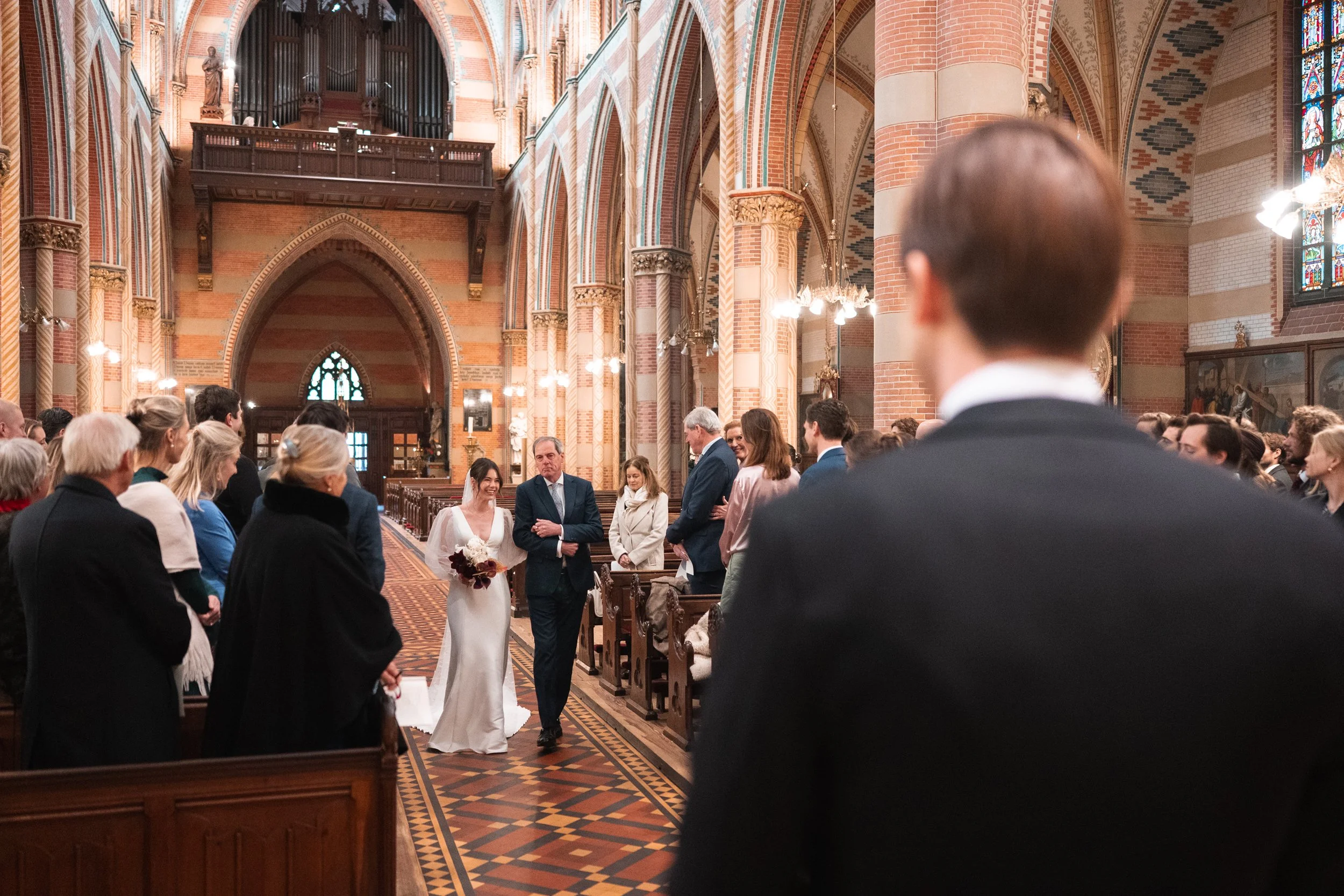 A wedding ceremony inside a church with stained glass windows and brick walls, showing the bride walking down the aisle towards the groom and officiant, surrounded by guests seated in pews.