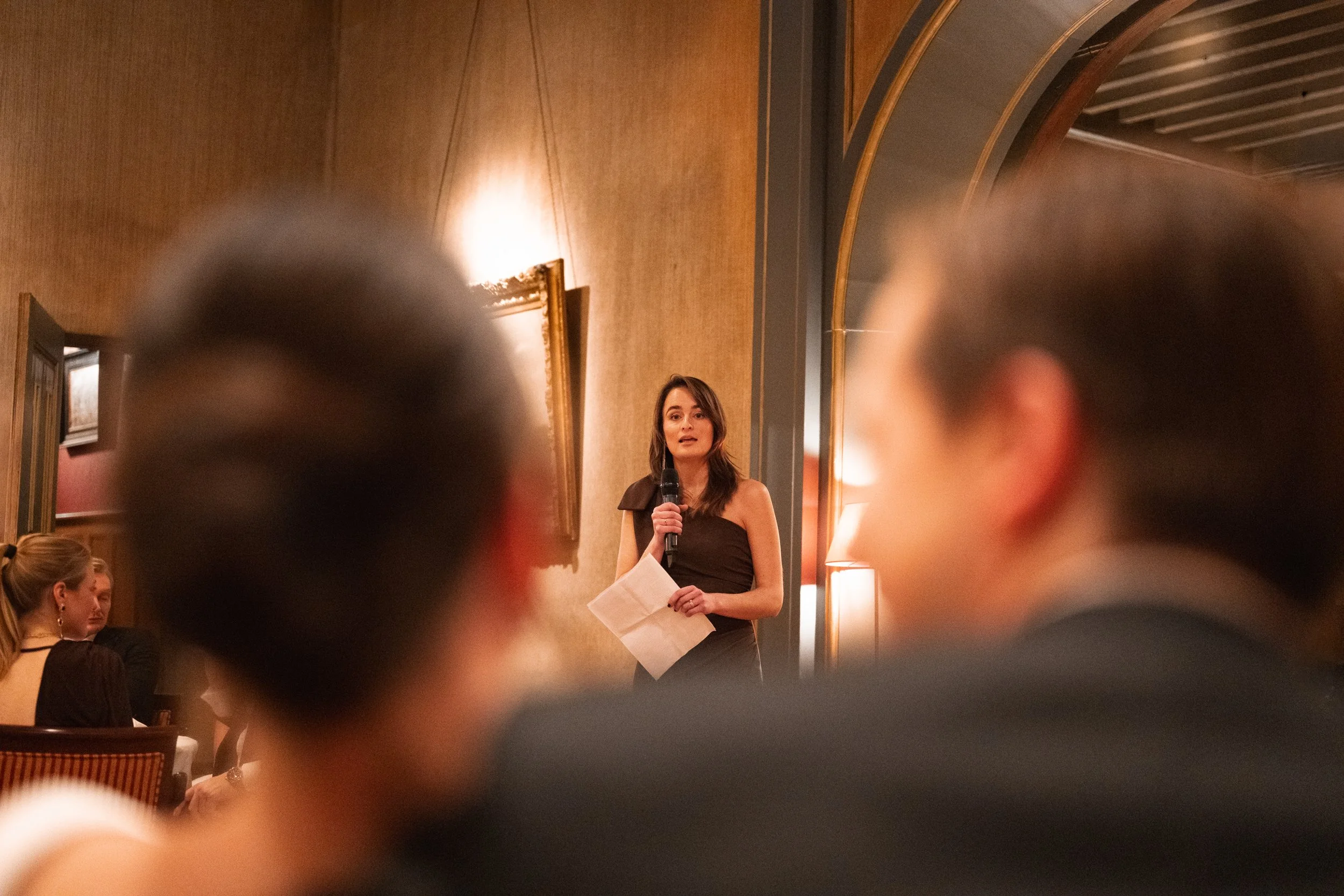 A woman in a black dress speaking into a microphone during an event, with blurred figures of audience members in the foreground and warm-toned decor in the background.