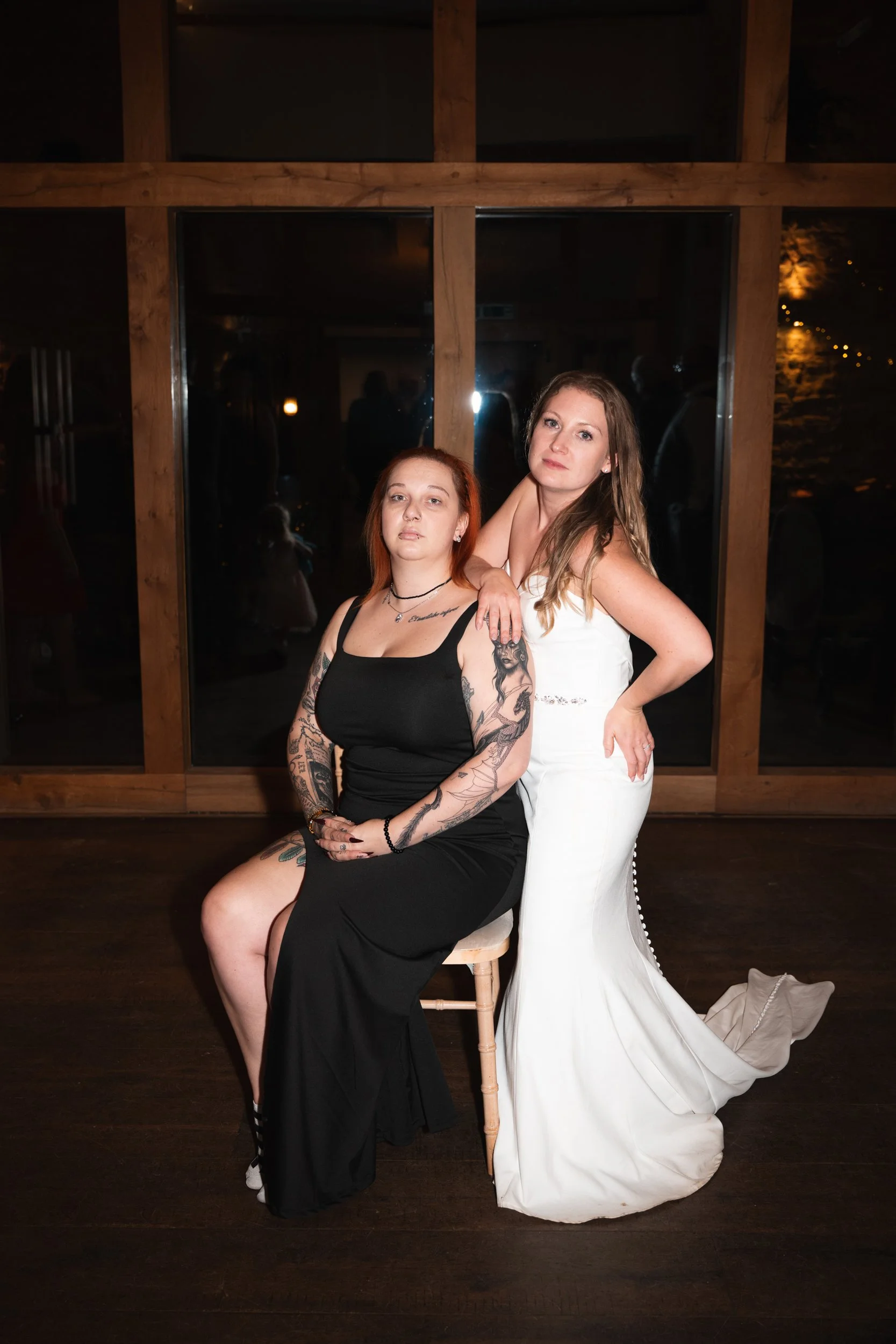 Two women pose together at a nighttime indoor event, one sitting on a stool wearing a black dress and tattoos, the other standing behind her in a white dress, against a wooden frame window background.