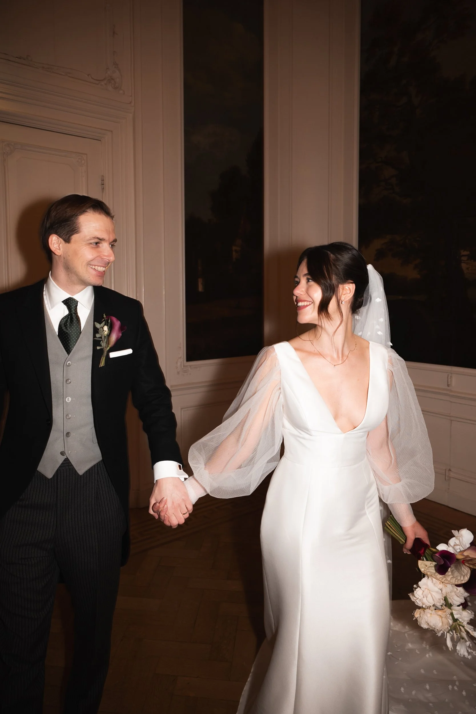 A bride and groom holding hands and smiling at each other indoors during their wedding reception, with the bride wearing a white gown with sheer puffy sleeves and a veil, and the groom wearing a black suit with a gray vest and a patterned tie.