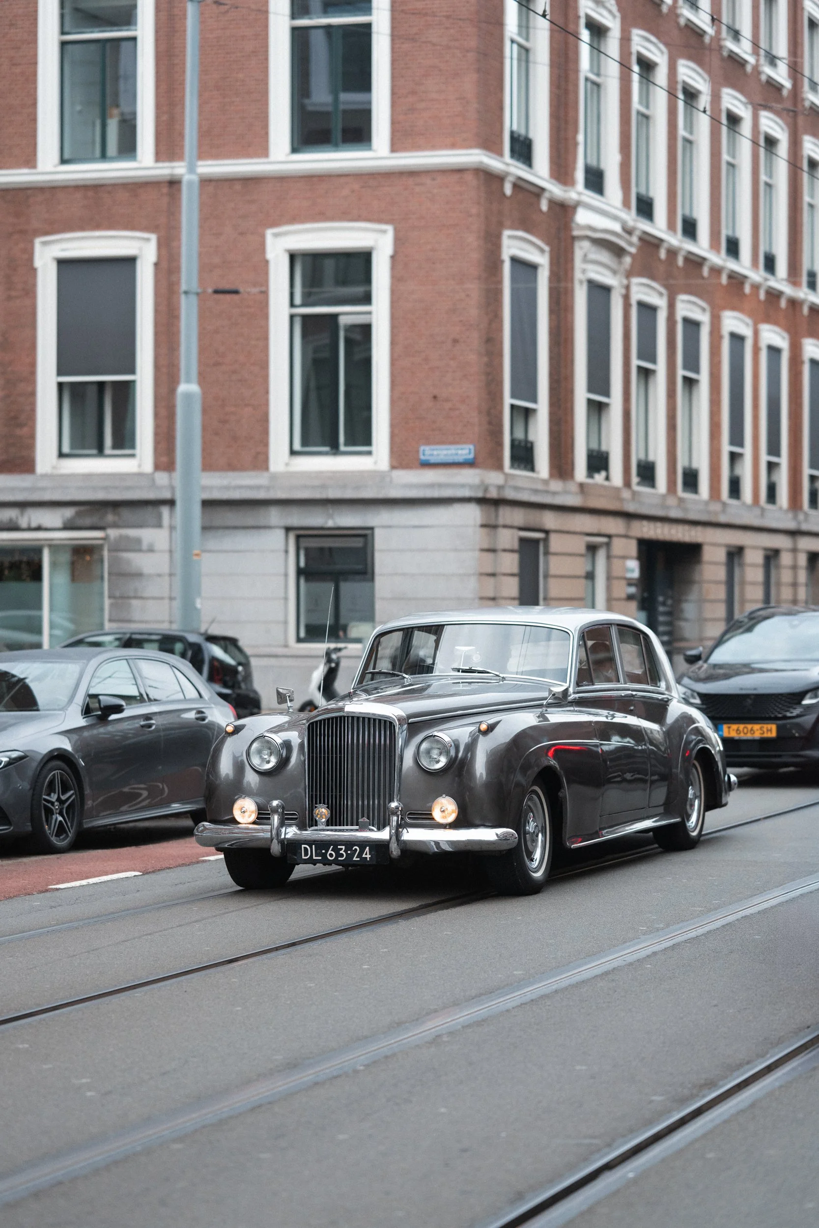 A vintage black luxury car driving on city street with brick building and modern parked cars in background.