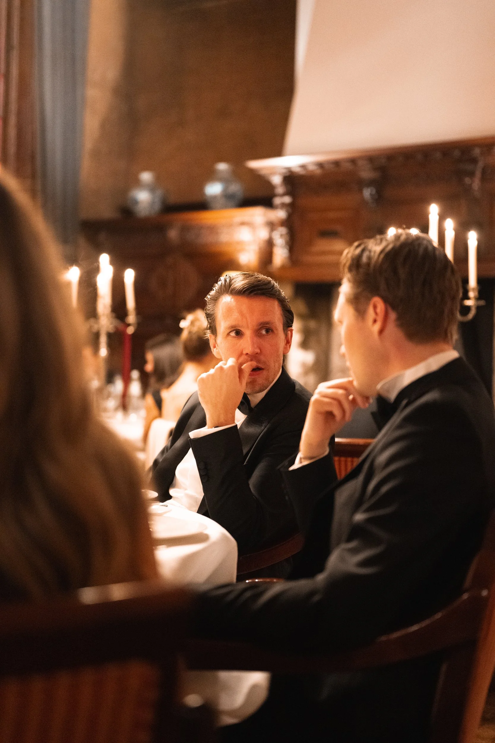 Two men in tuxedos sitting at a dinner table, engaged in conversation, in a dimly lit, elegant restaurant with candles and wooden decor.