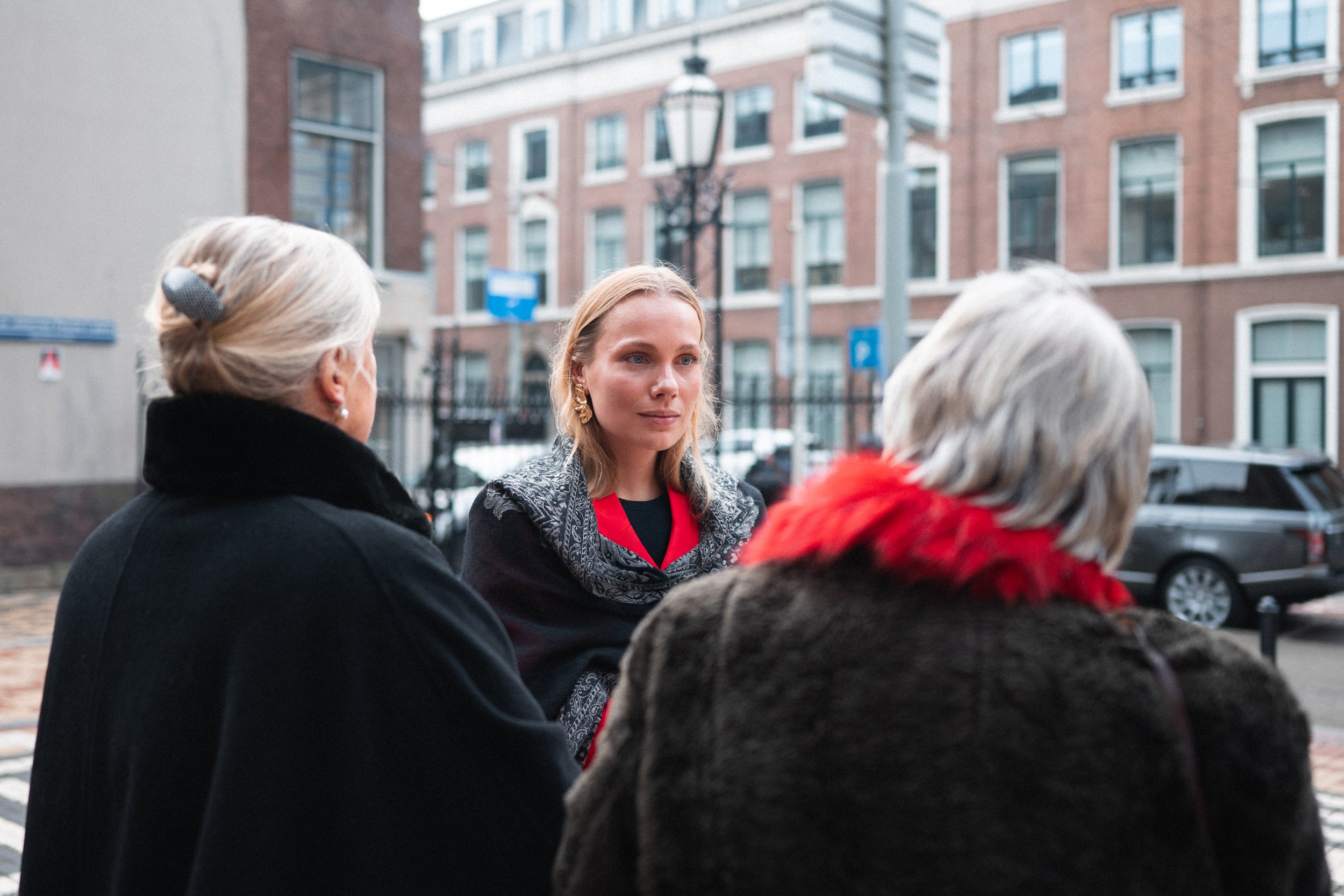 Three women standing outdoors in an urban environment, having a conversation. One woman with blonde hair, wearing makeup and earrings, faces the camera, while two other women with gray hair, one with a hair clip and red fur collar, face away from the