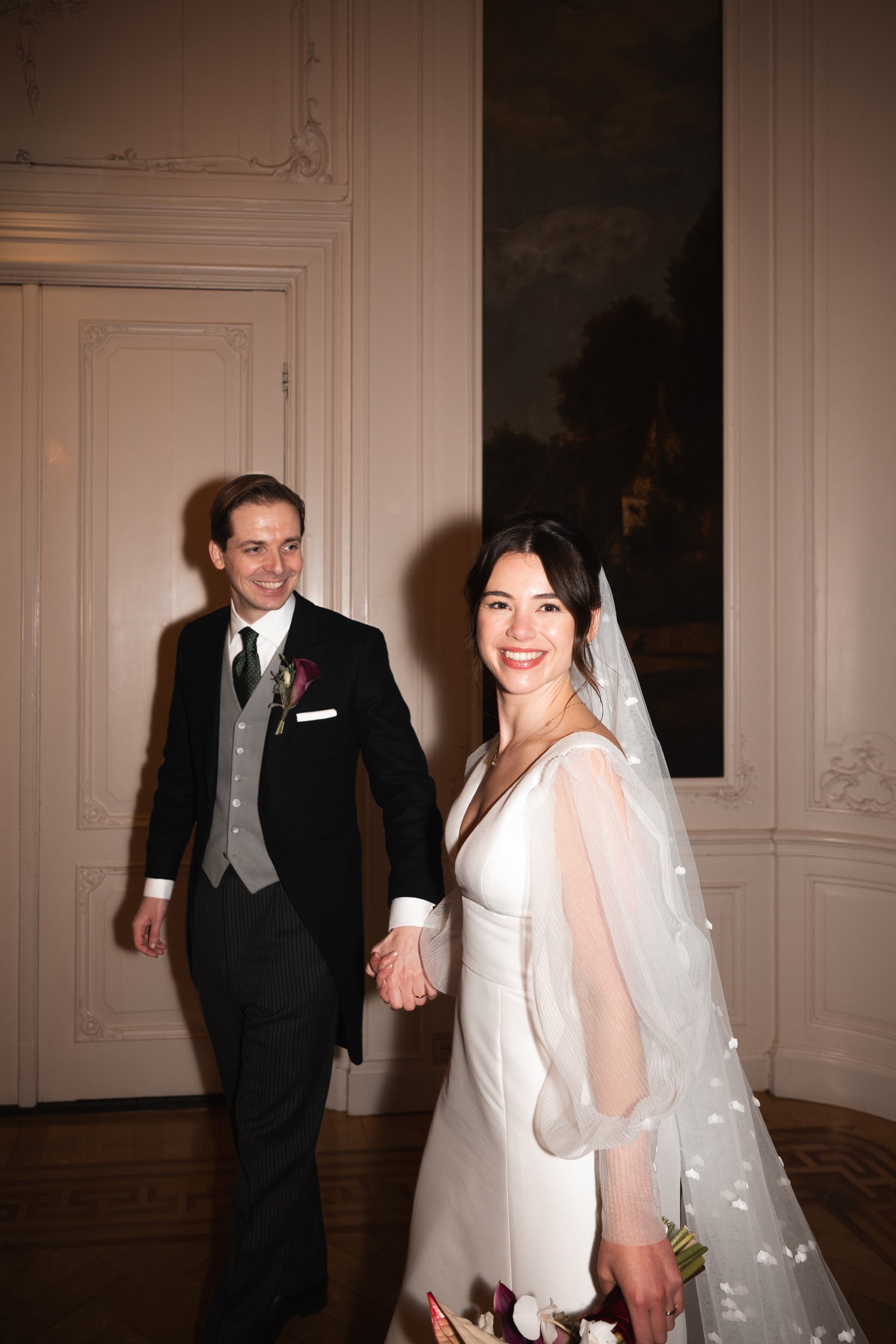 A bride and groom holding hands and smiling at each other indoors. The bride is wearing a white wedding dress and veil, holding a bouquet. The groom is dressed in a formal black suit with a grey vest and tie.