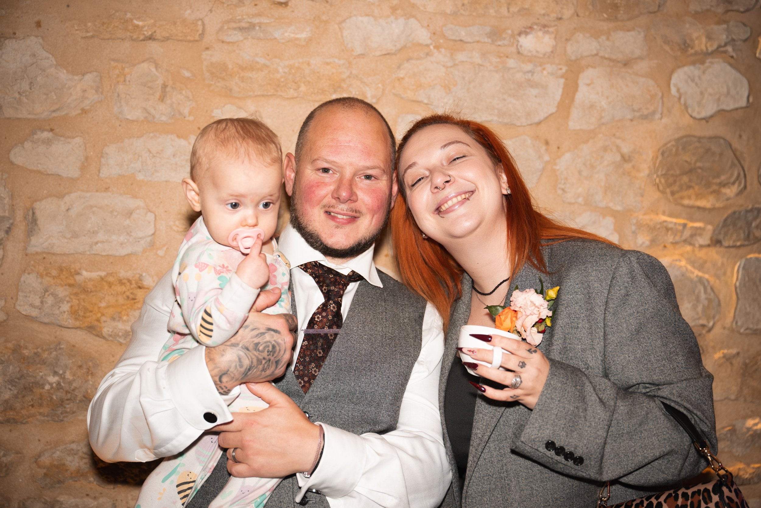 A smiling family of three posing indoors against a stone wall. The man is holding a young girl with a pacifier, and the woman is holding a cup, wearing a flower boutonniere, and smiling with her eyes closed.
