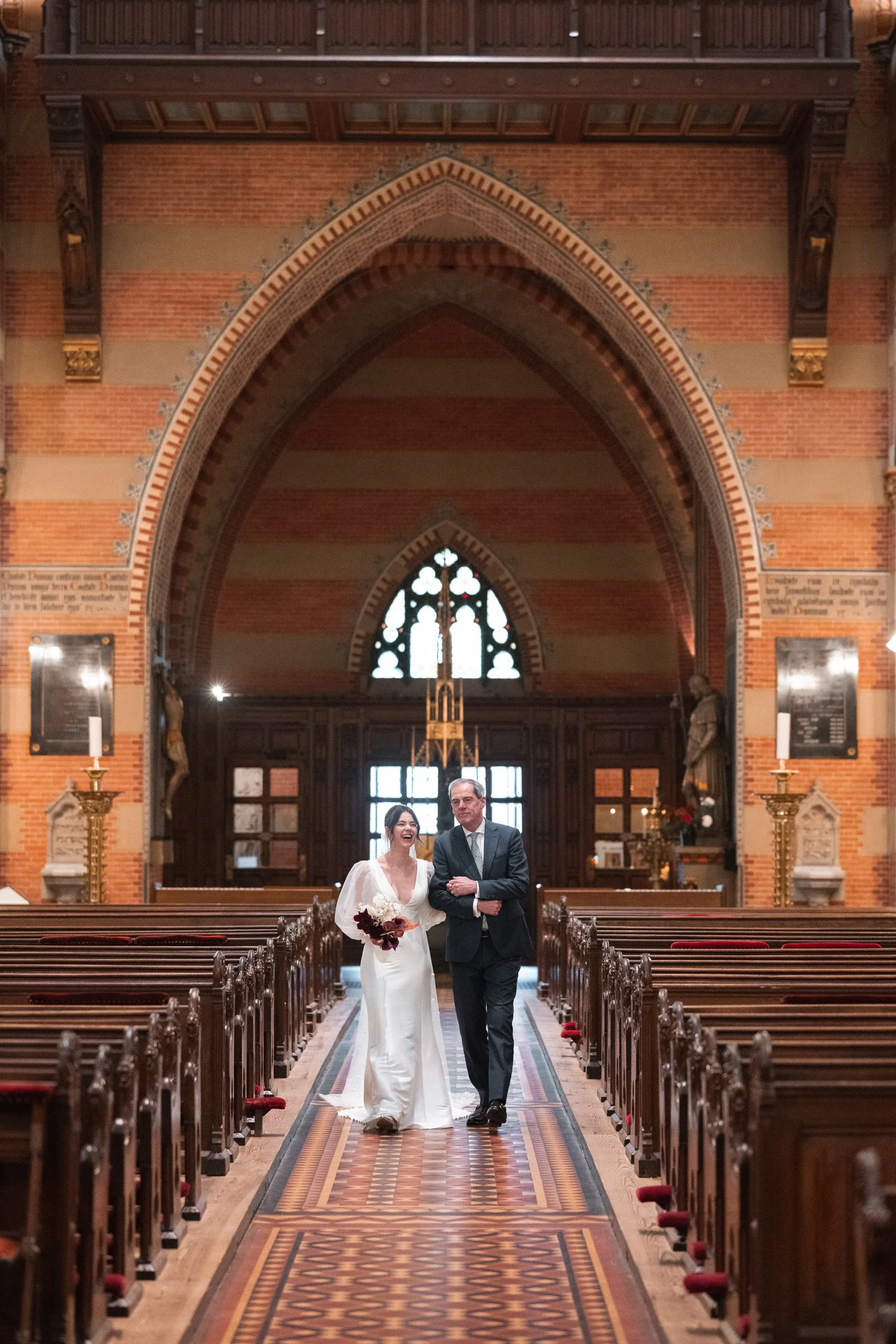 A bride in a white wedding dress walking down the aisle with an older man in a suit inside a church with high arched ceilings, stained glass windows, and wooden pews.