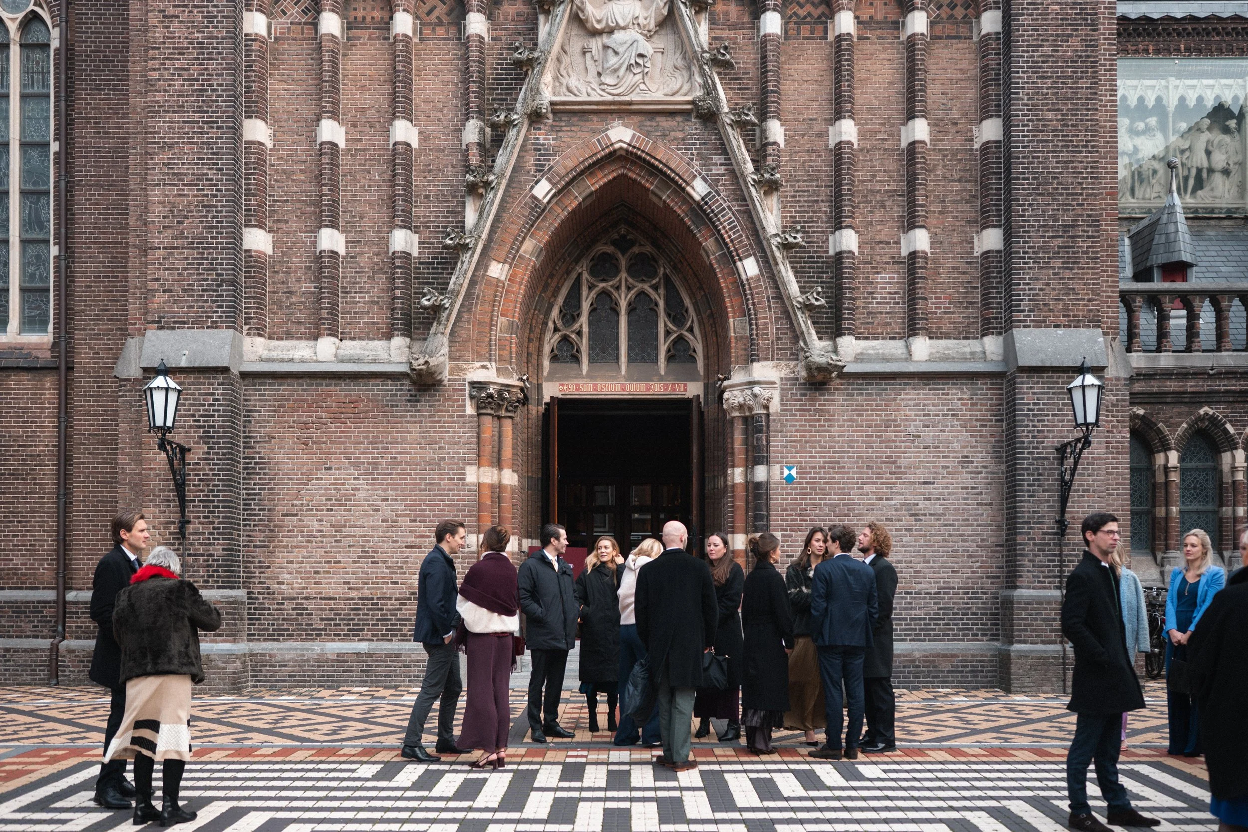 A group of people dressed in formal and semi-formal attire standing and talking in front of a large brick church entrance with Gothic architectural details, situated on a patterned sidewalk.