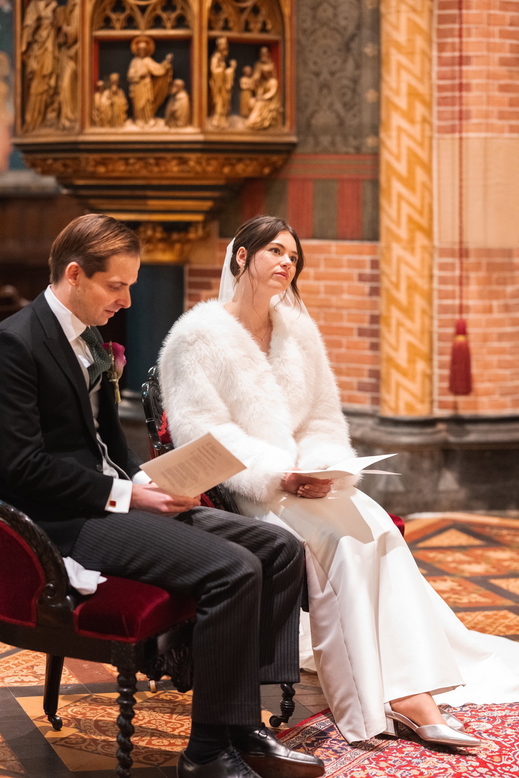 A bride in a white wedding dress and fur coat sitting next to a groom in a black suit inside a church, both reading from papers during a wedding ceremony.