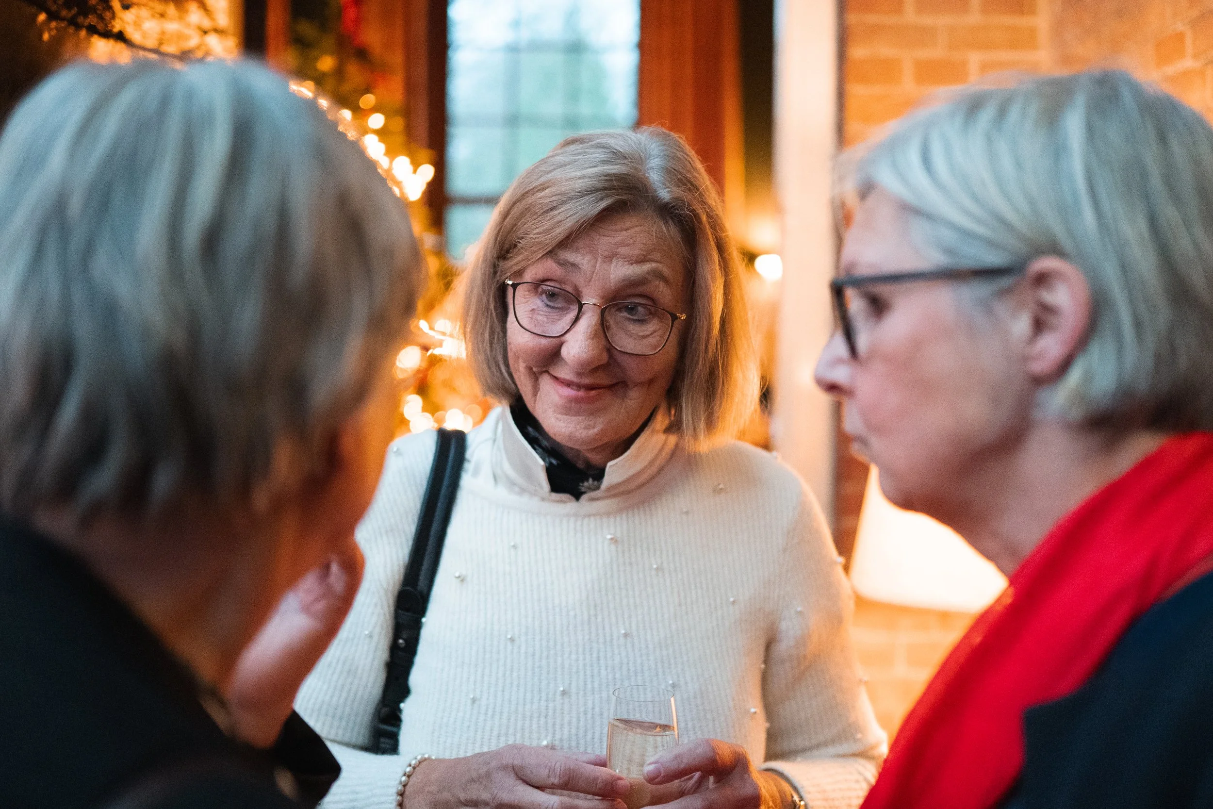 Three elderly women conversing at a social gathering, one woman holding a champagne glass. The background features warm lighting and a brick wall.