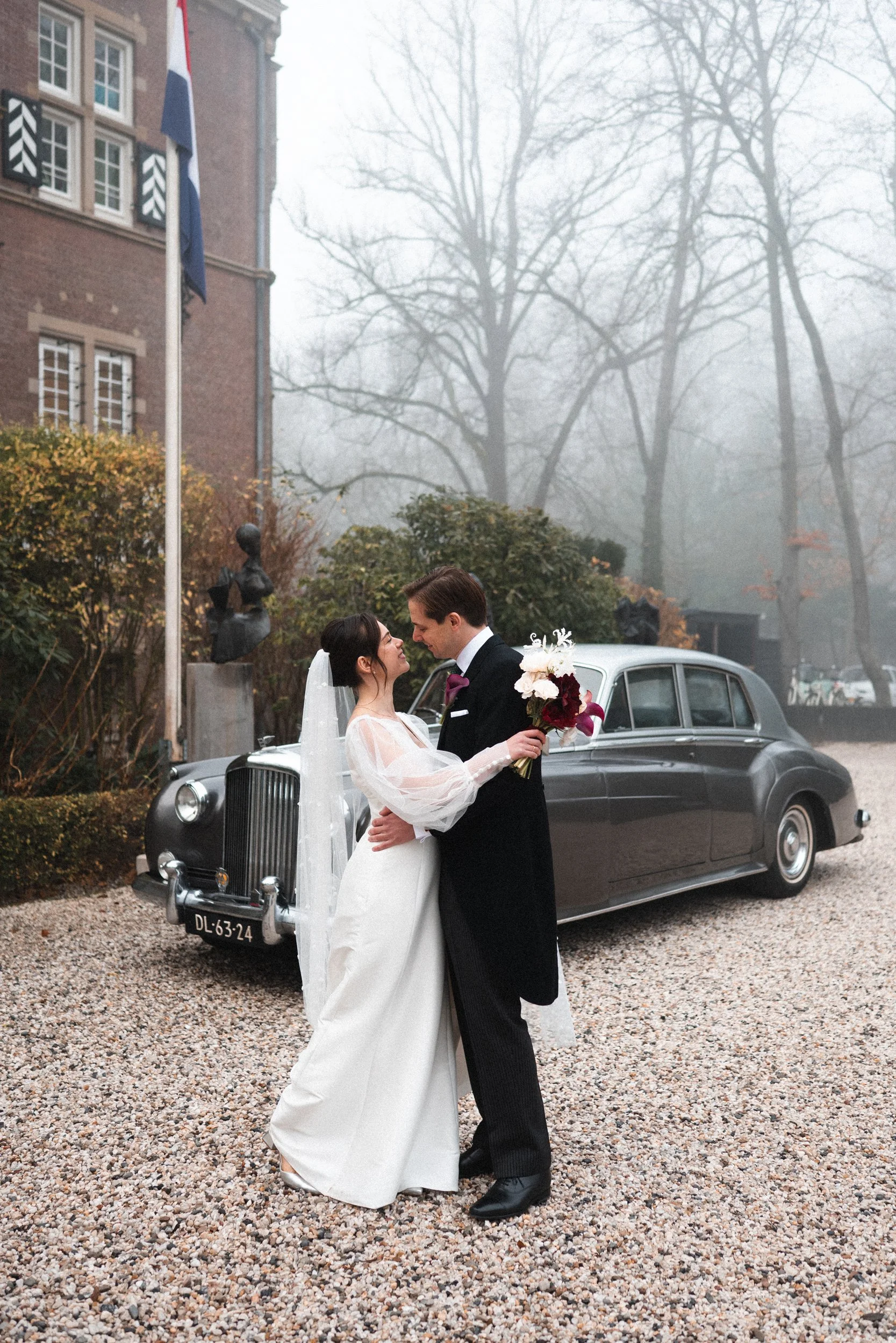A bride and groom sharing a romantic moment outdoors on their wedding day, standing in front of a vintage black car with a foggy, leafless background and a building with flags.
