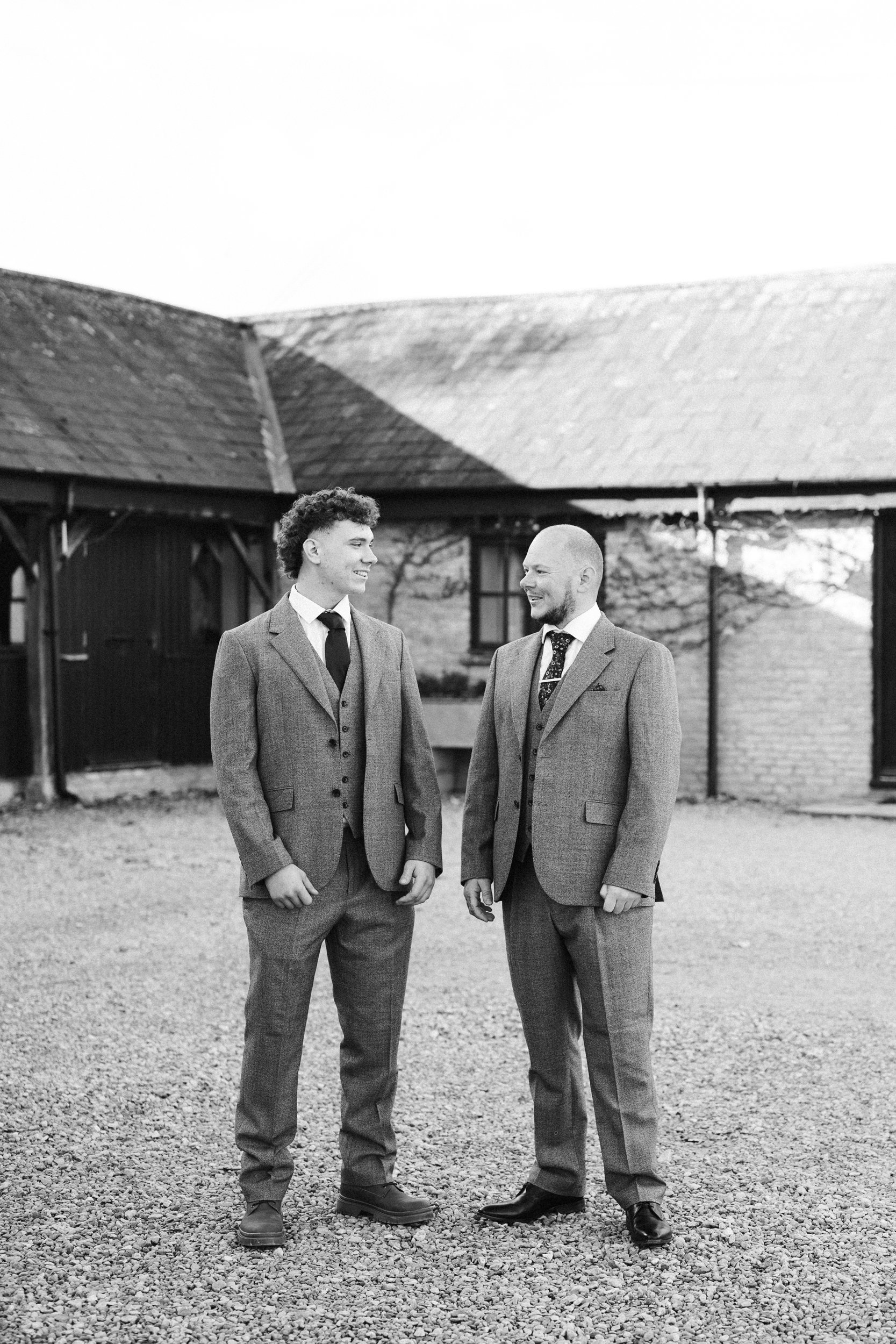 Two men in suits standing outdoors and smiling at each other, in front of a rustic building with a sloped roof in black and white.
