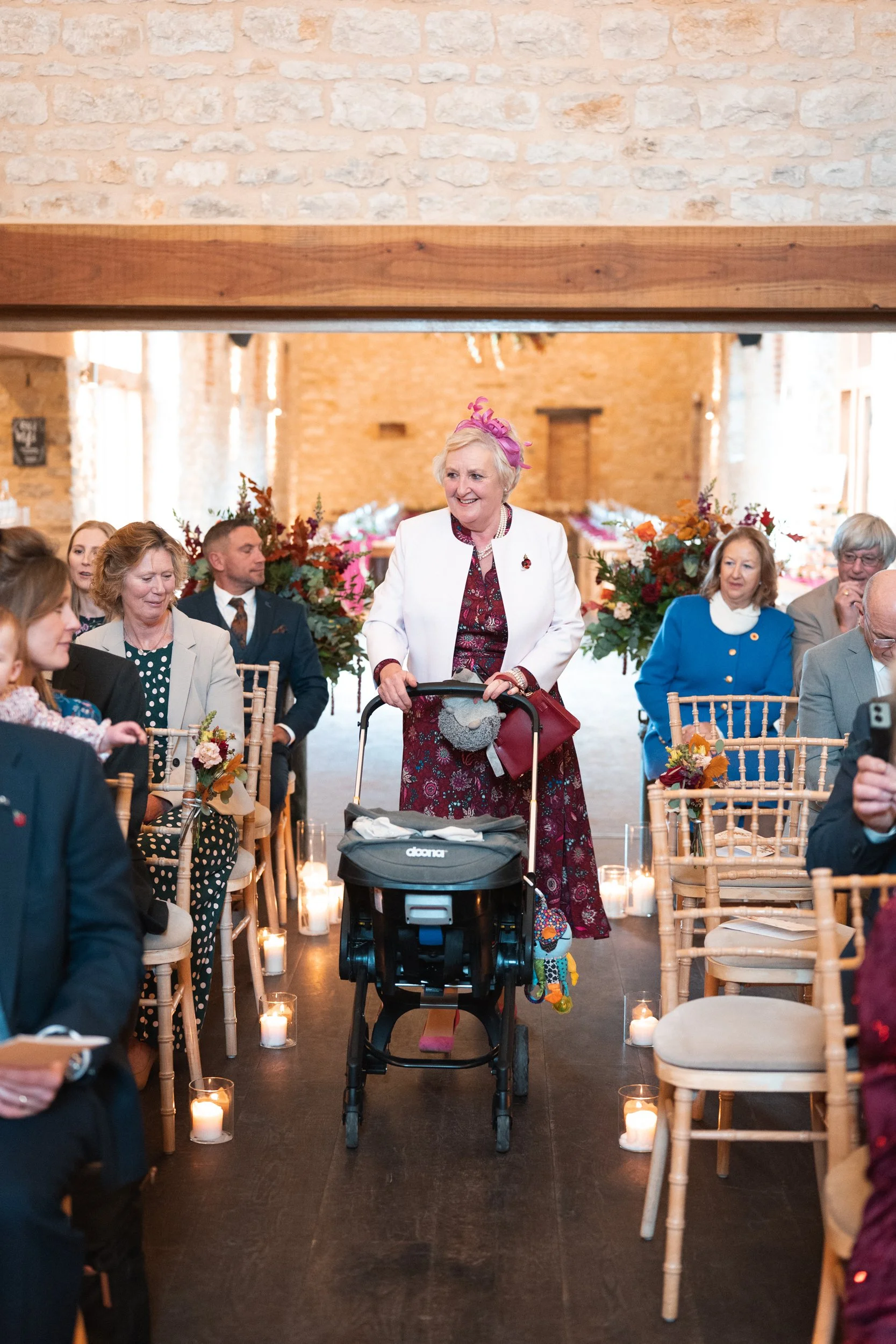 An elderly woman with a pink fascinator hat, white blazer, and maroon dress, smiling and pushing a stroller through a decorated indoor event space with seated guests, floral arrangements, and lit candles.
